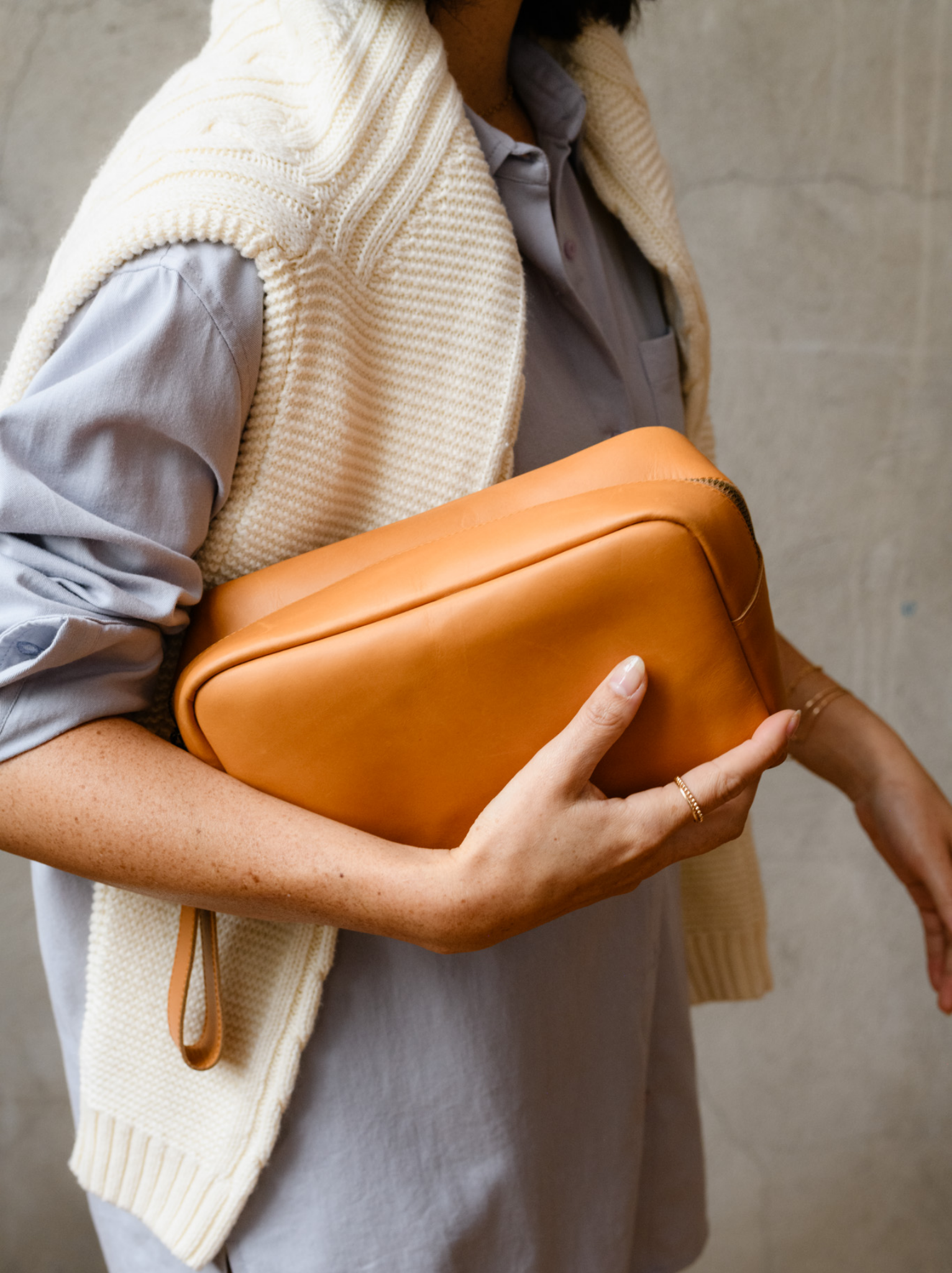 A woman holding a cognac full grain pull up leather travel case with a hang strap. #Color_Cognac
