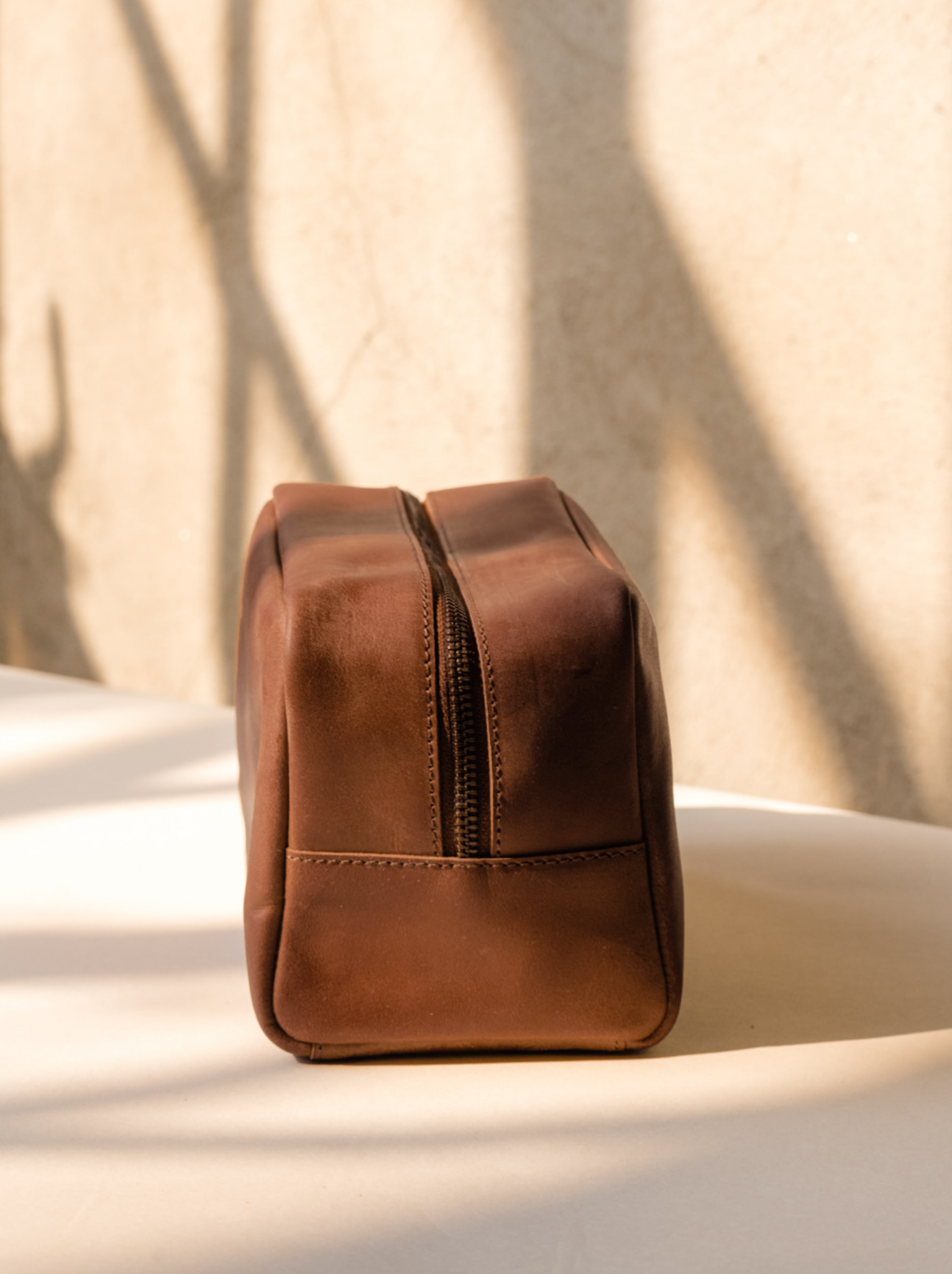 Zipper detail of brown leather dopp kit on light colored table with shadows. #Color_Whiskey