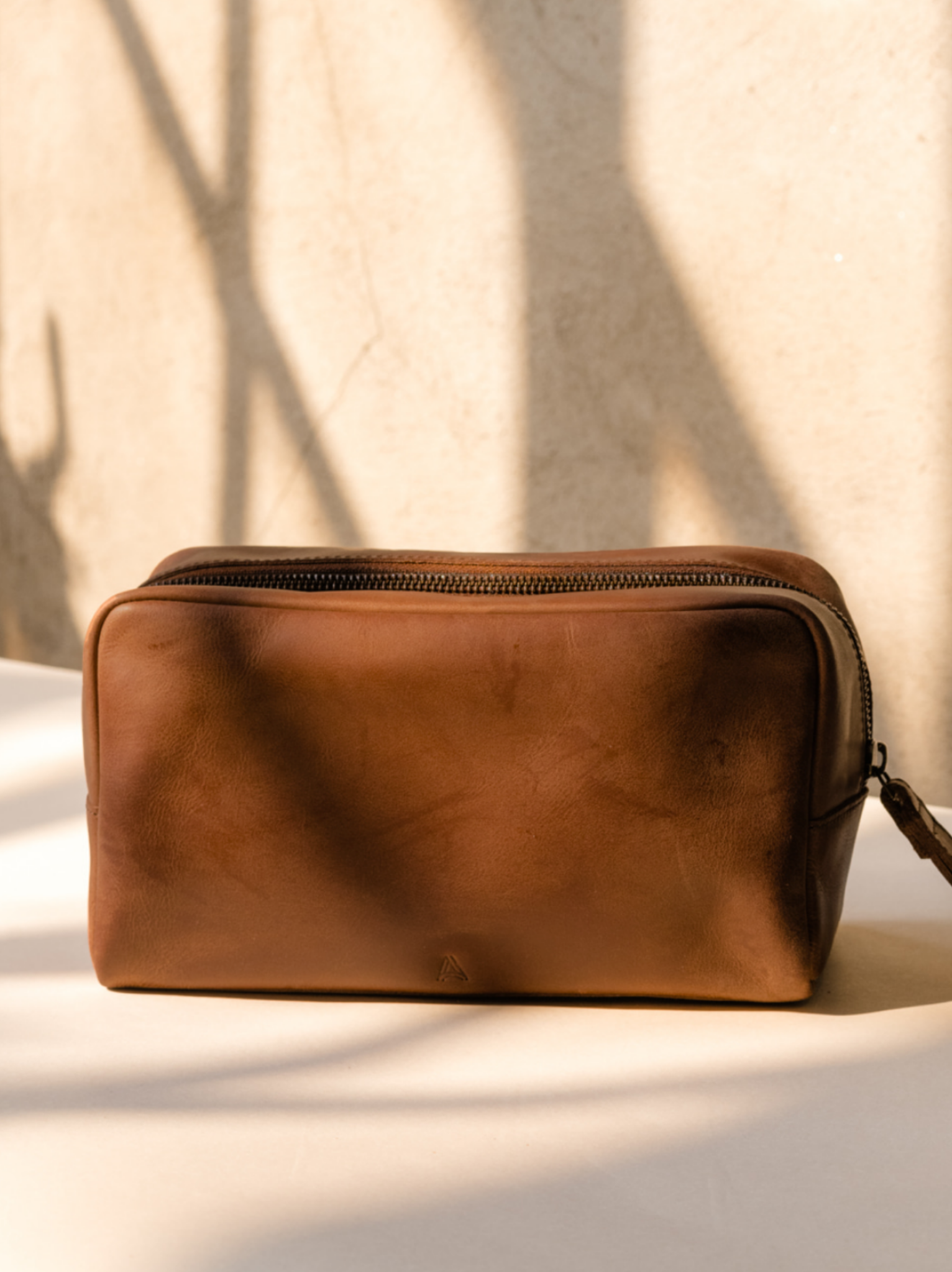Side profile of brown leather dopp kit against neutral background. #Color_Whiskey