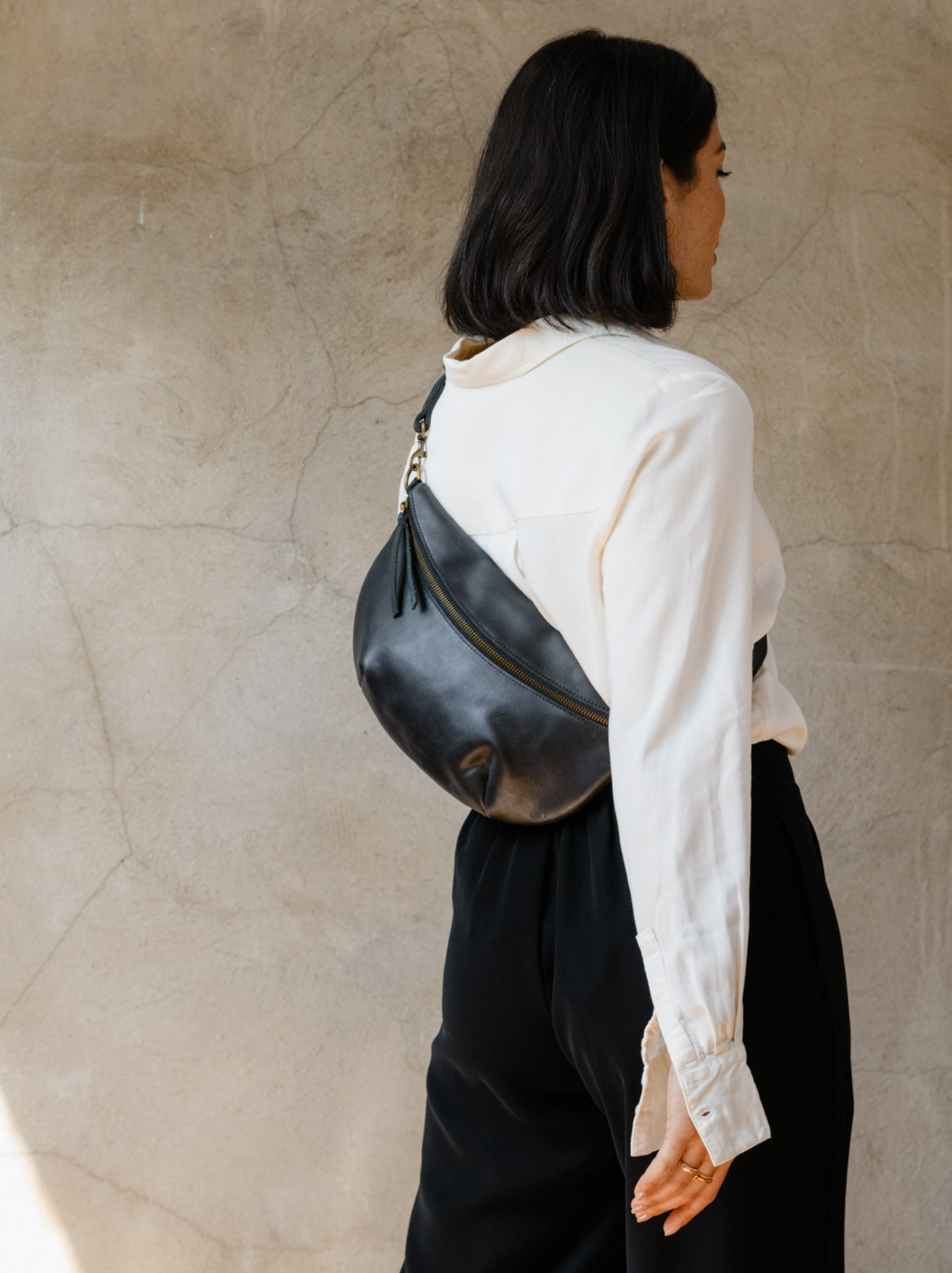 Woman wearing a black leather belt bag across her back, in a white blouse and black trousers, against a concrete wall. #Color_Black