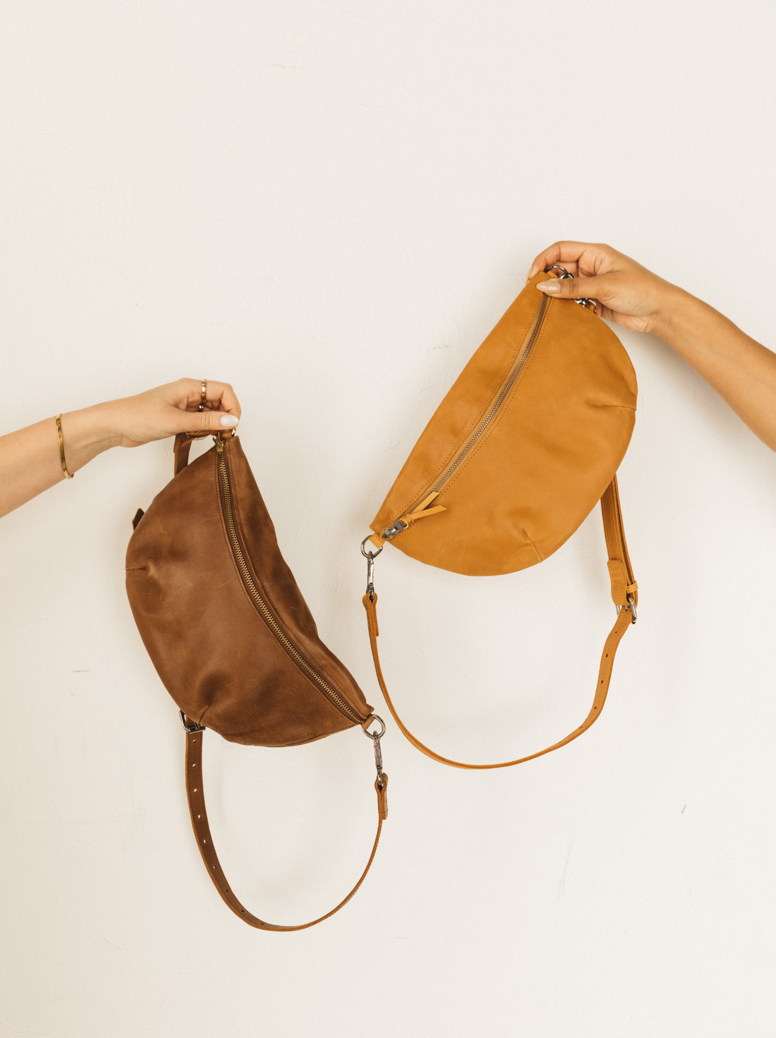 Women hands holding one brown leather belt bag and one cognac leather belt bag with crossbody straps hanging against white background. Core Colors #Color_Cognac