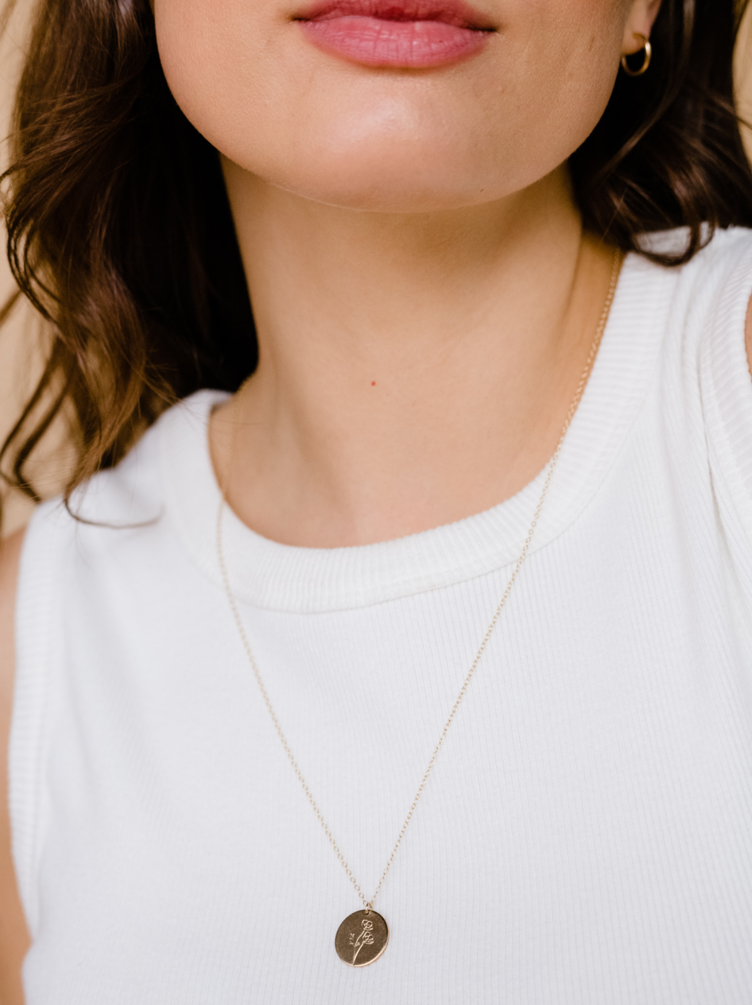 A woman wearing a 24 inch necklace with a round tag pendant with an engraved birth month flower against a white tank top.