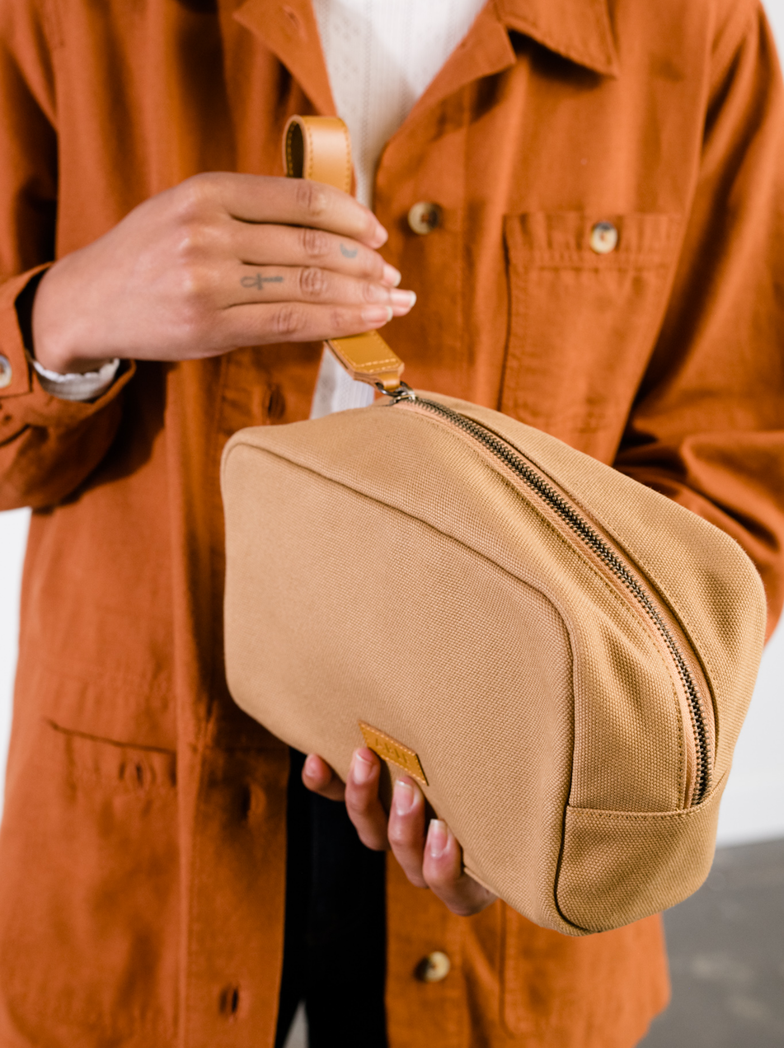 Woman unzipping a tan canvas toiletry bag using the cognac leather strap with an orange jacket