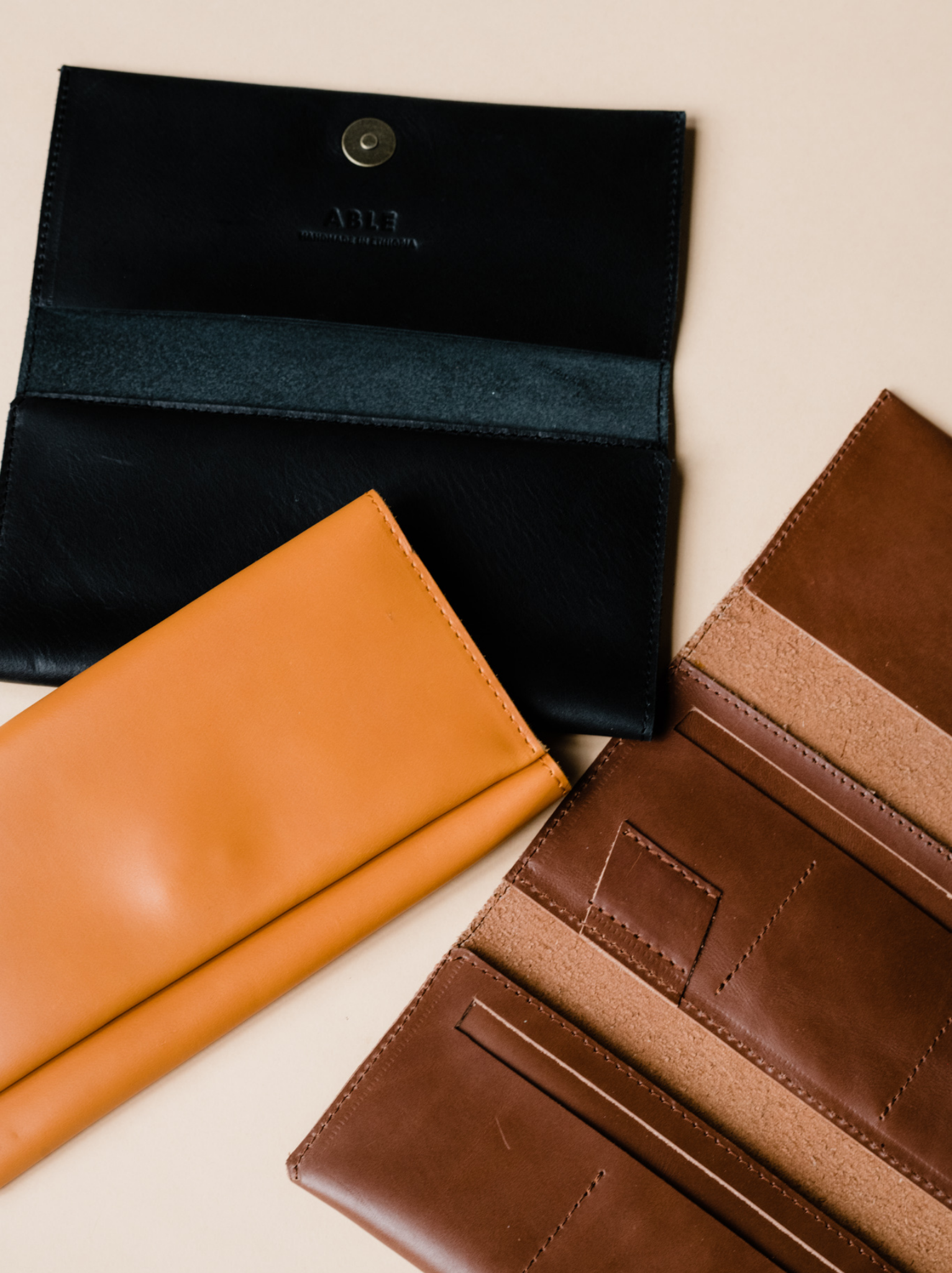Three leather wallets, brown, orange cognac, and black, lying next to each with one closed, one partially open and one completely open on neutral background. #Color_Cognac