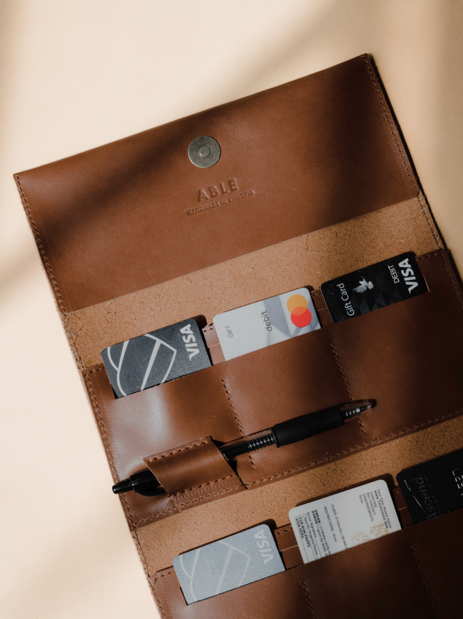 Opened trifold brown leather wallet with credit cards in slots and pen in pen holder with magnetic closure on neutral background. #Color_Whiskey