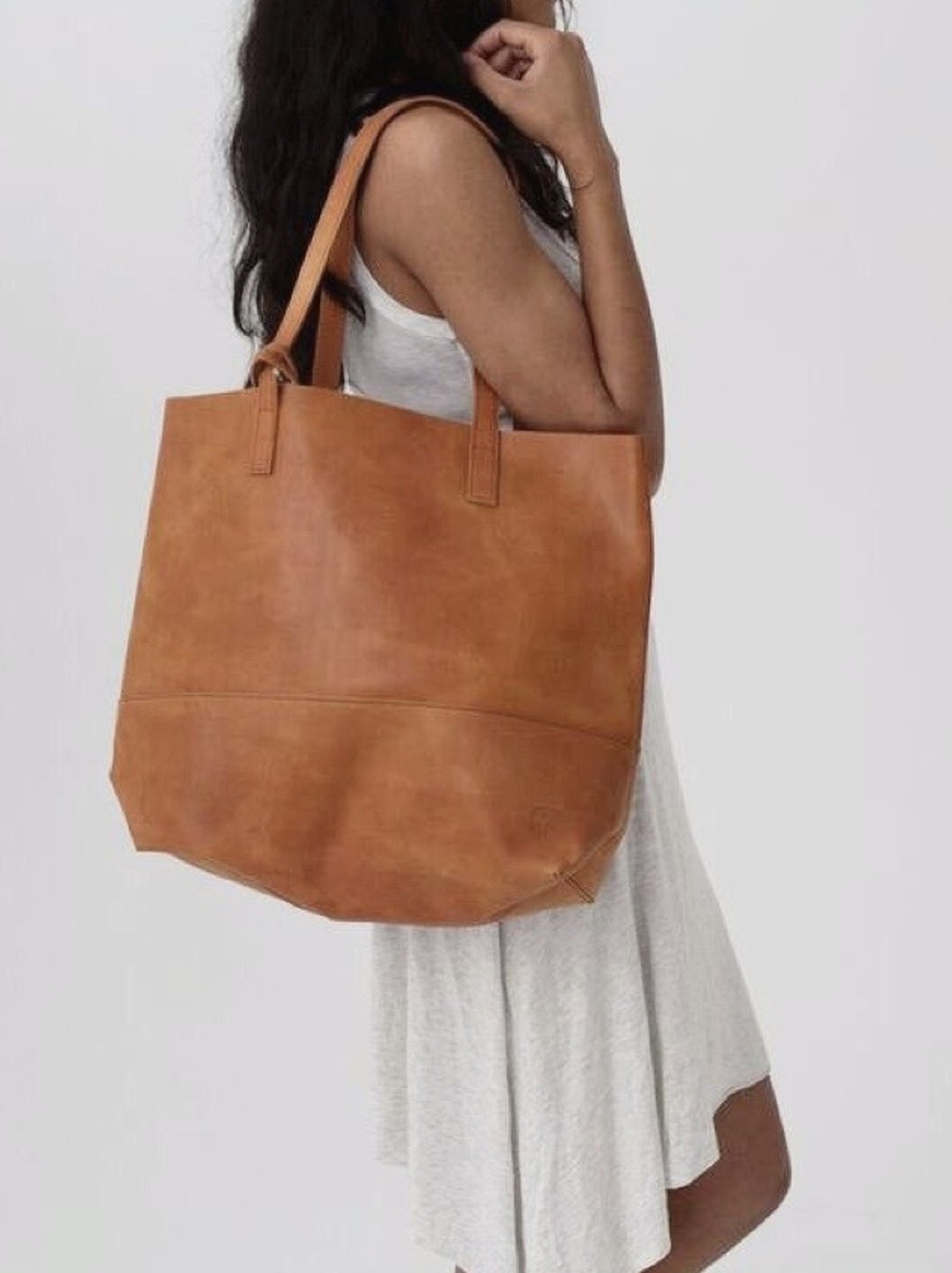 Woman carrying a cognac leather tote on her shoulder wearing a white dress against a white background. #Color_Cognac