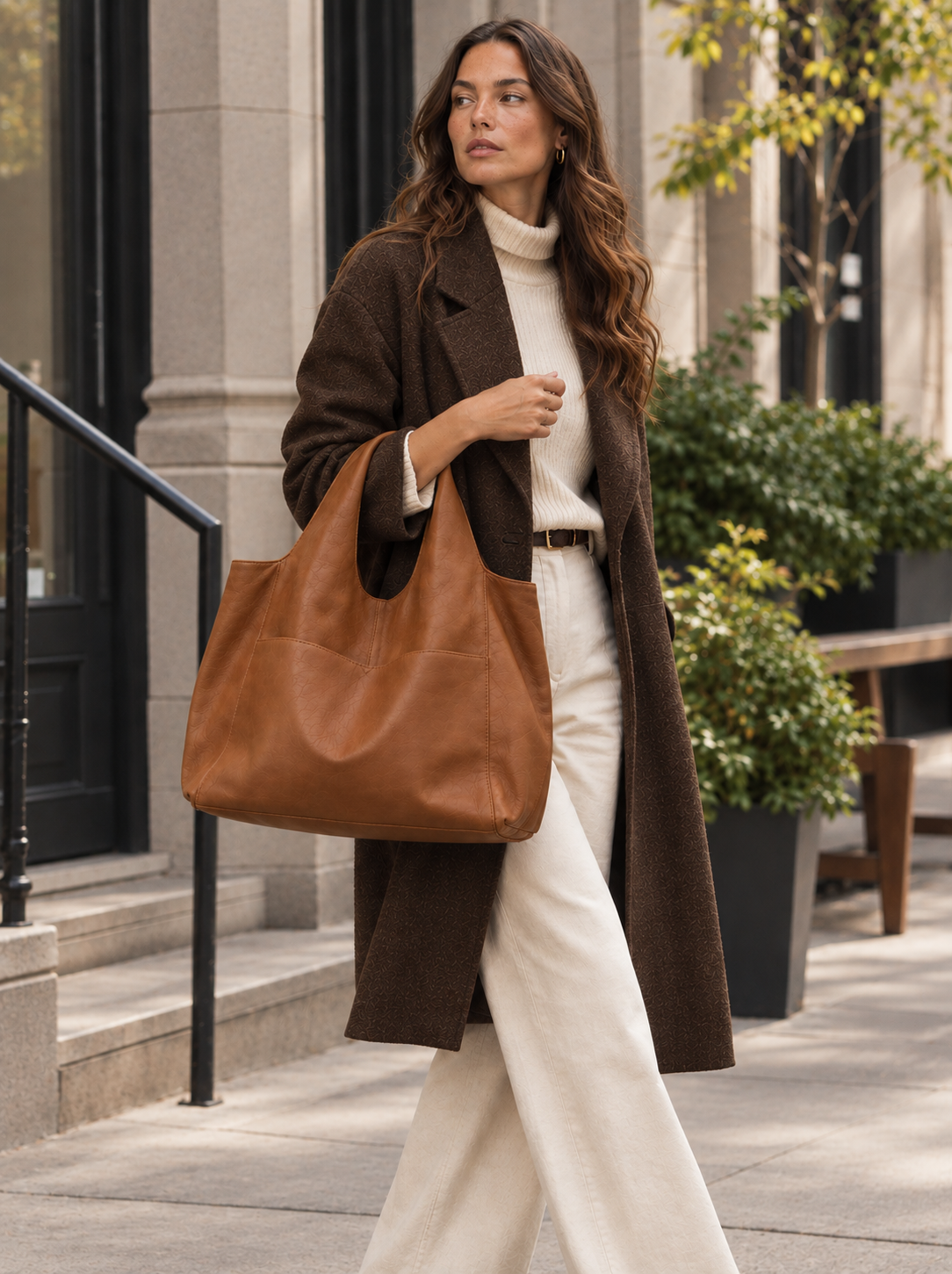 A woman walking on a city sidewalk carrying a large brown leather tote bag. She is wearing a brown coat, a cream turtleneck sweater, and cream wide-leg pants. #Color_Chestnut