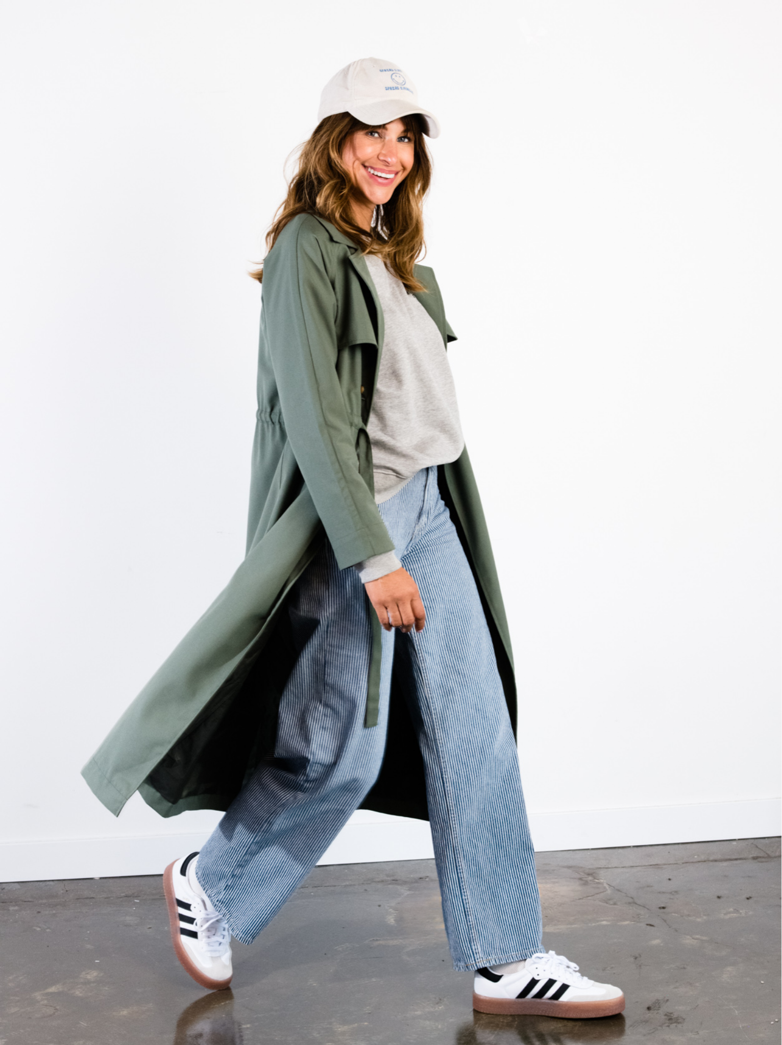 A woman walking indoors with long sleeve green trench coat, sweatshirt, jeans, and sneakers. #Color_Moss