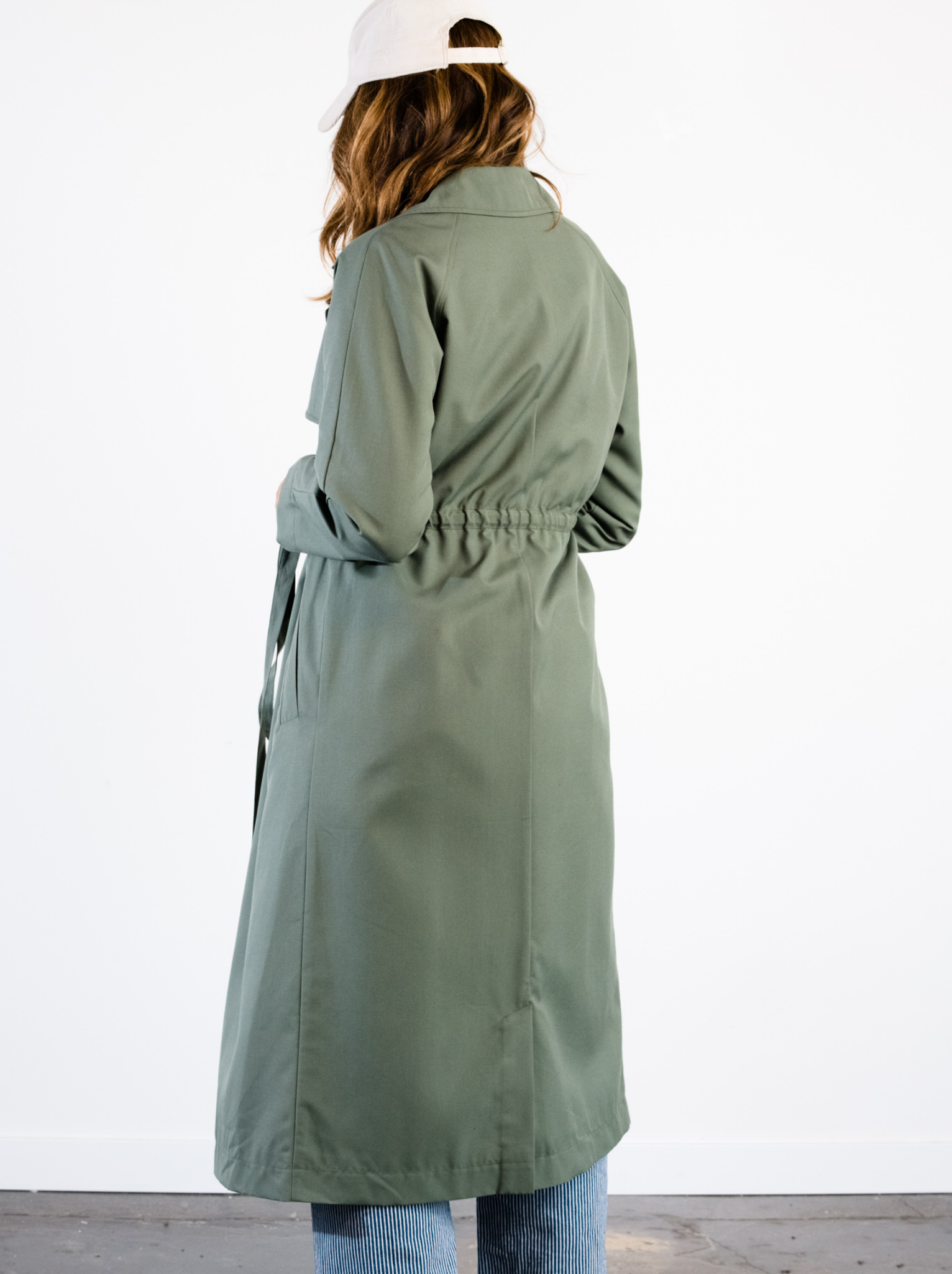 Back of a woman wearing a green trench coat against a white wall. #Color_Moss