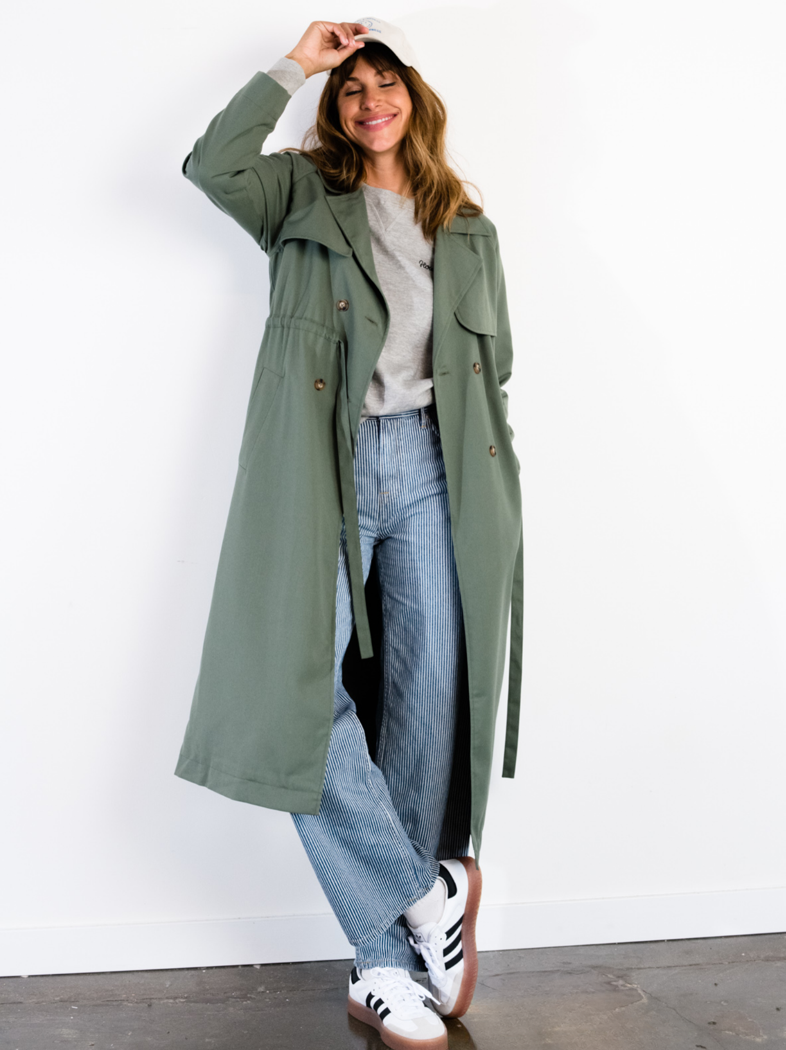 A woman leaning against a white wall wearing a green trench coat with long sleeves and hand in one pocket. #Color_Moss