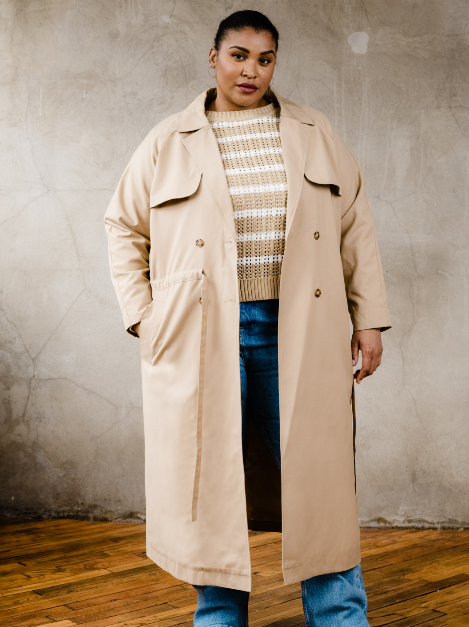 Woman wearing unbelted tan trench coat with hand in one pocket, tan stripe sweater and denim jeans indoors on wooden floor with neutral background. #Color_Brown Sugar
