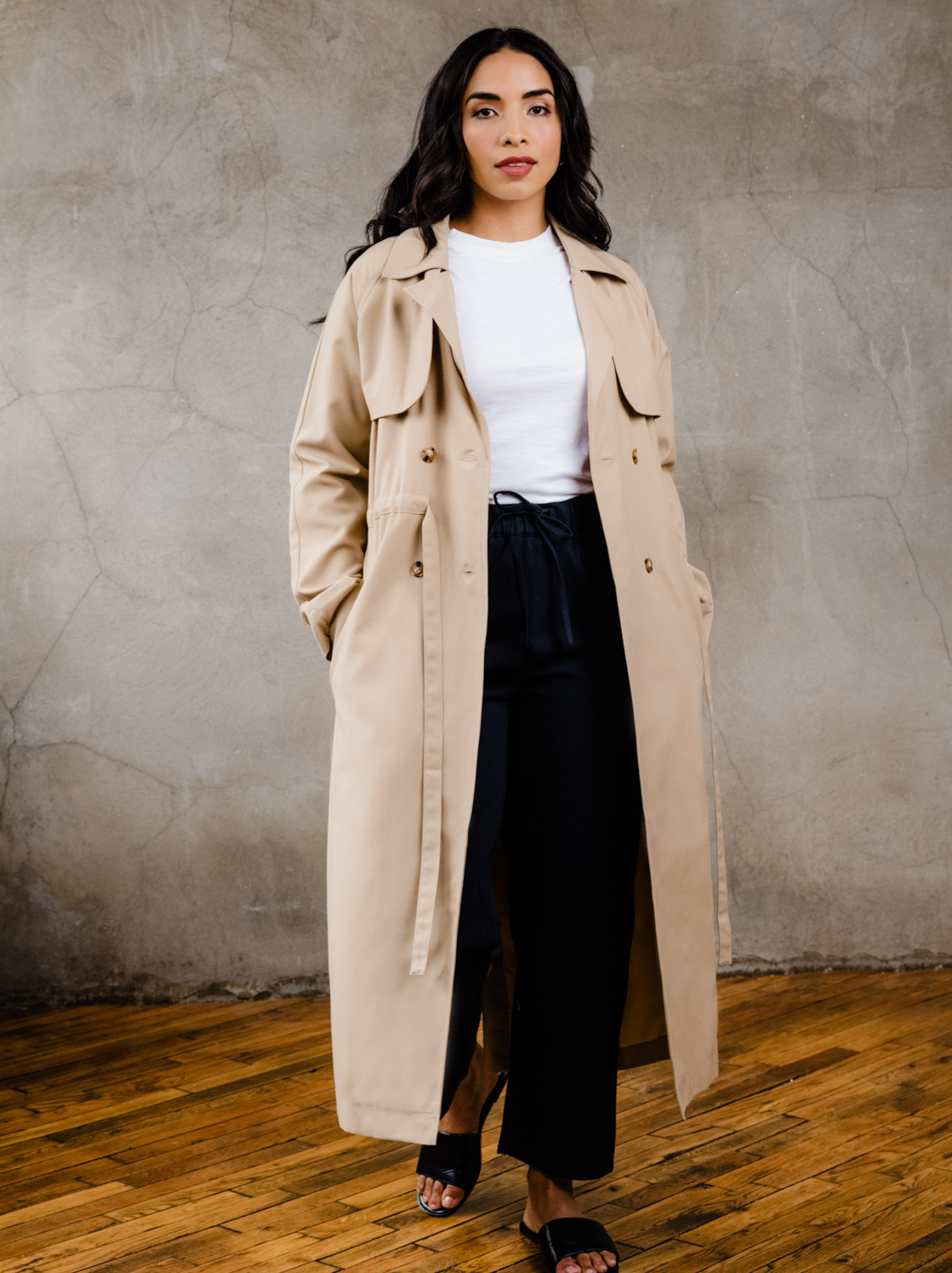 Woman wearing unbelted beige trench coat, hands in both pockets, with white t-shirt, black pants, and black sandals indoors on wooden floor with neutral background. #Color_Brown Sugar