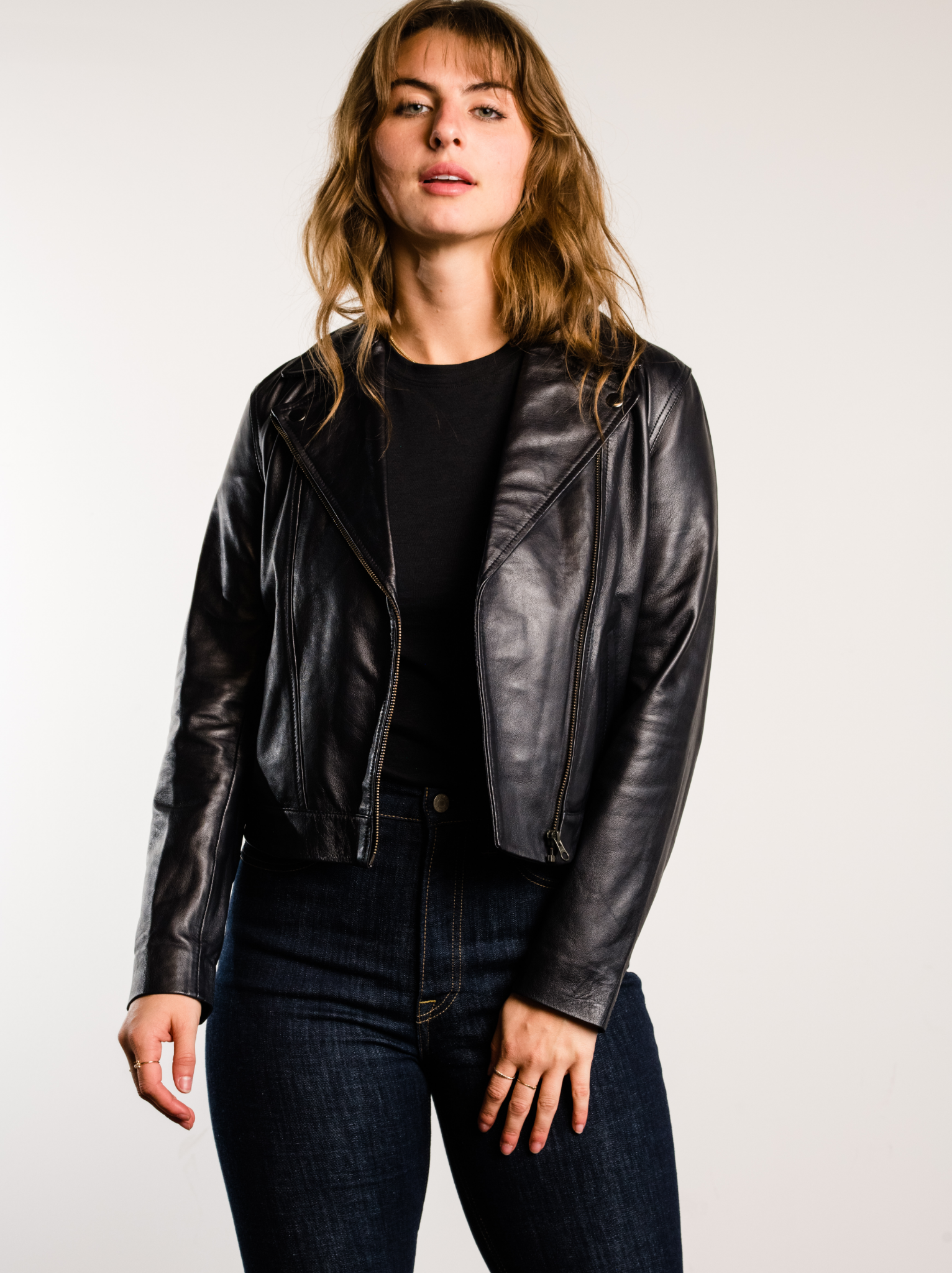 A woman wearing a black leather jacket with black tank and dark jeans against a white background. #Color_Black