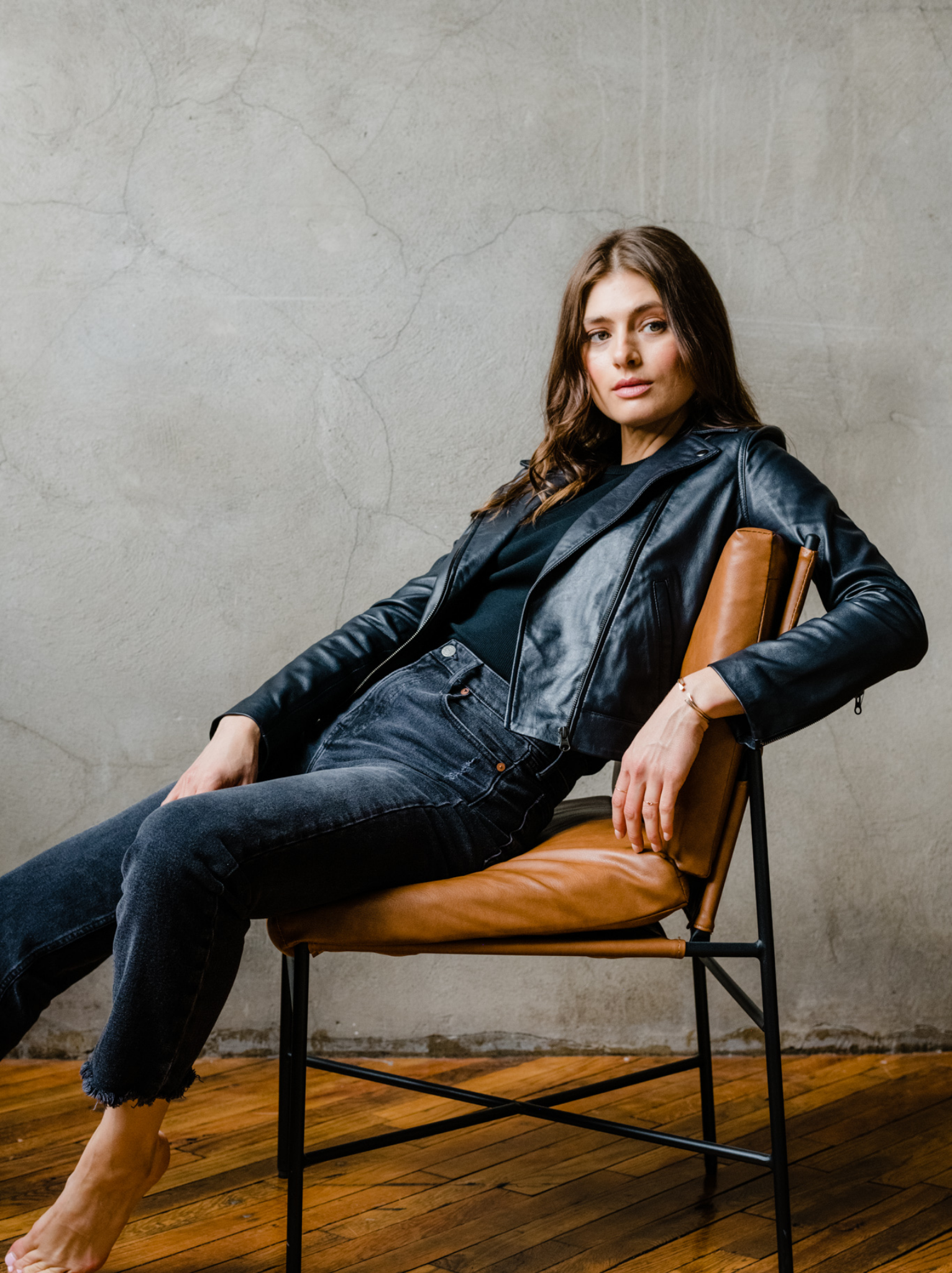 A woman sitting in a chair wearing a black leather jacket, black tank, and black jeans. #Color_Black