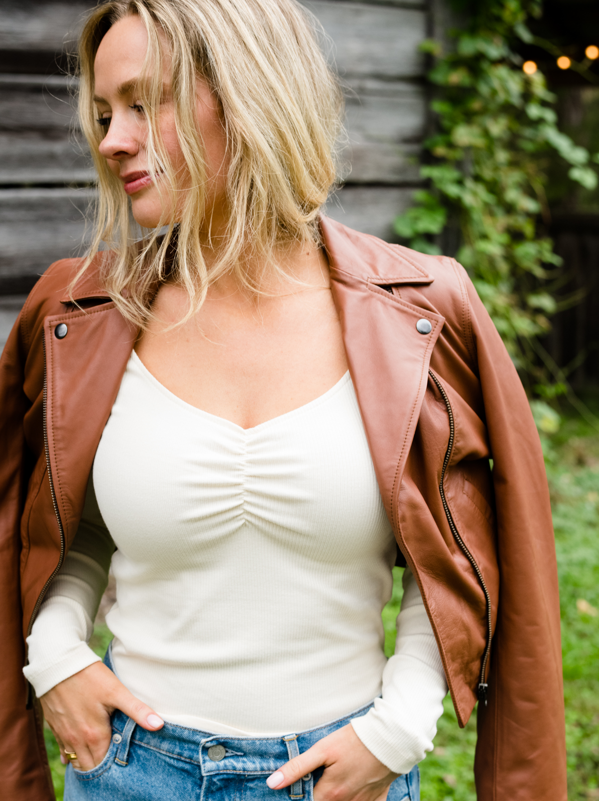A woman outside with her hands in her jean pockets and wearing a brown leather jacket on her shoulders. #Color_Whiskey