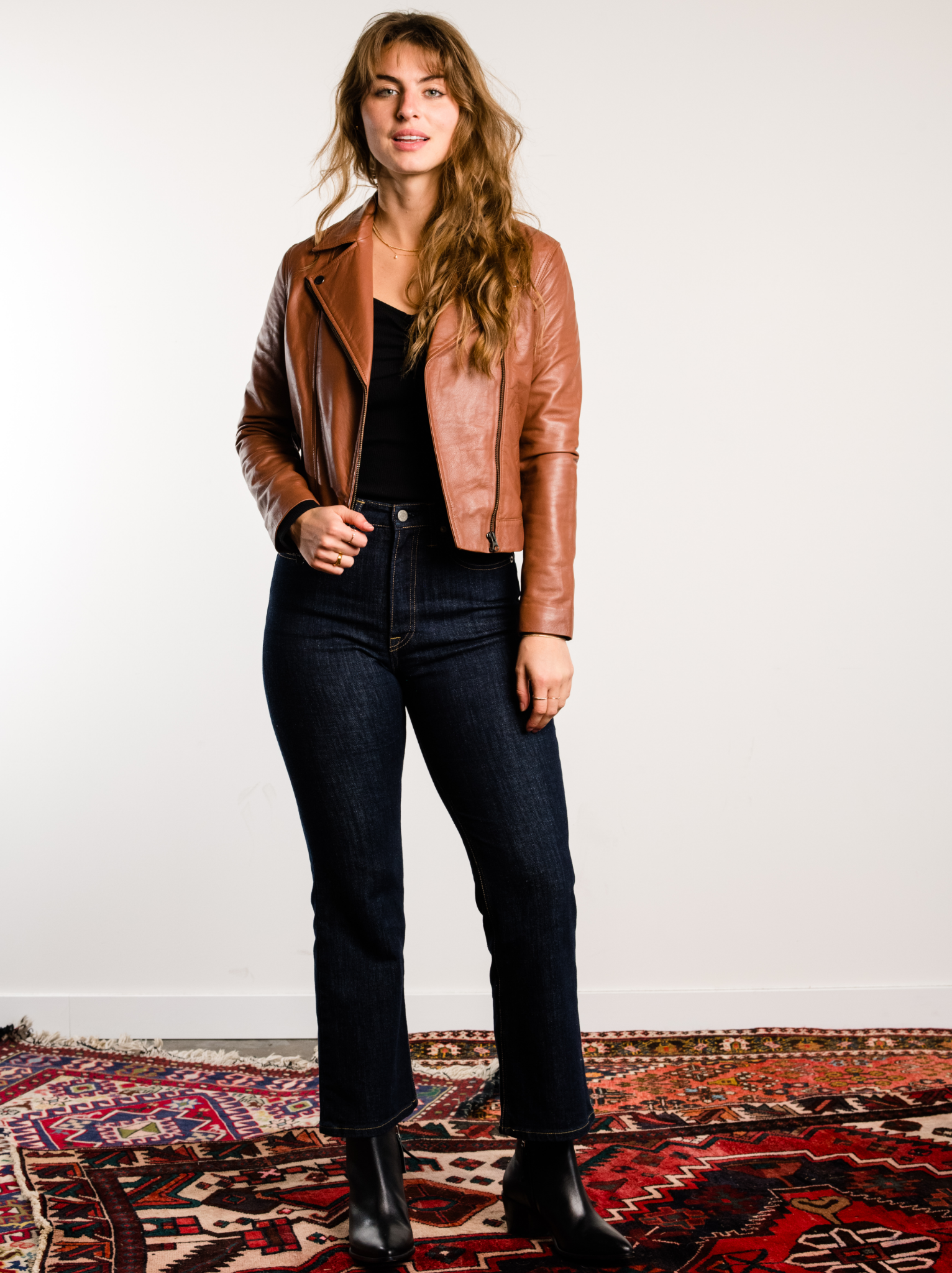 A woman wearing a brown leather jacket with black top, dark jeans, and black boots on colorful rugs against in front of a white wall. #Color_Whiskey