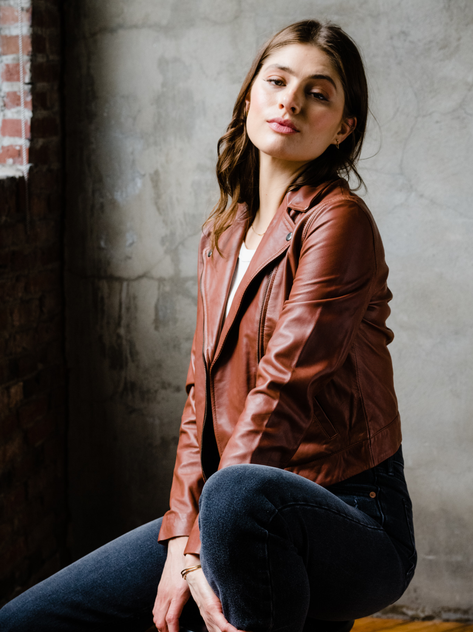 A woman sitting on a stool wearing a brown leather jacket and jeans indoors with a neutral background. #Color_Whiskey