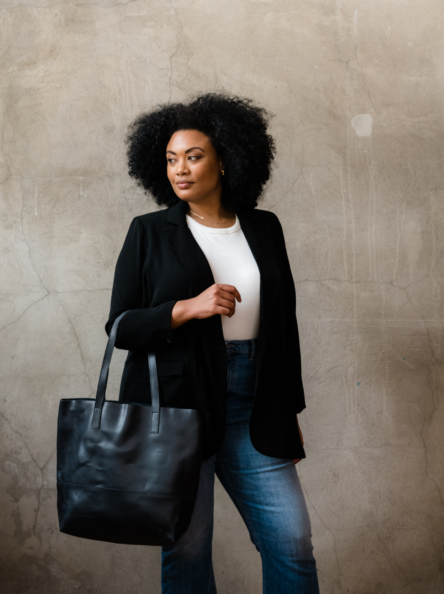 Woman holding a black leather tote by two straps on her forearm, wearing a black and jeans against a neutral background. #Color_Black