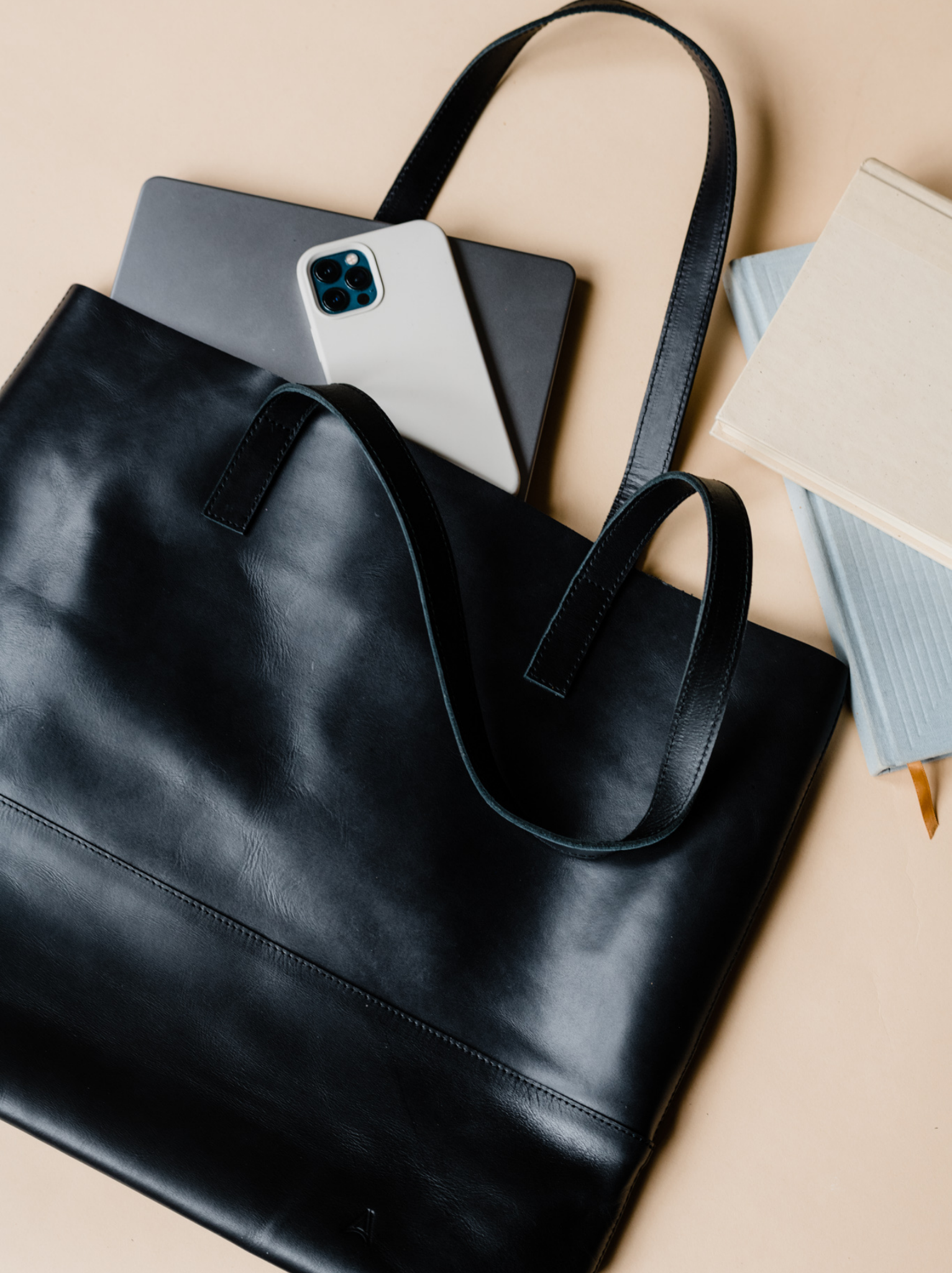 Black leather tote laying on a table with a laptop, phone, and books coming out of it. #Color_Black