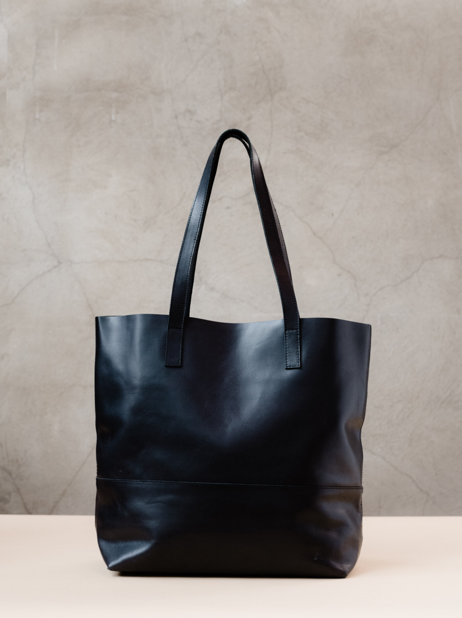 Black leather tote bag with two shoulder straps sitting on a tan surface with a neutral background. #Color_Black