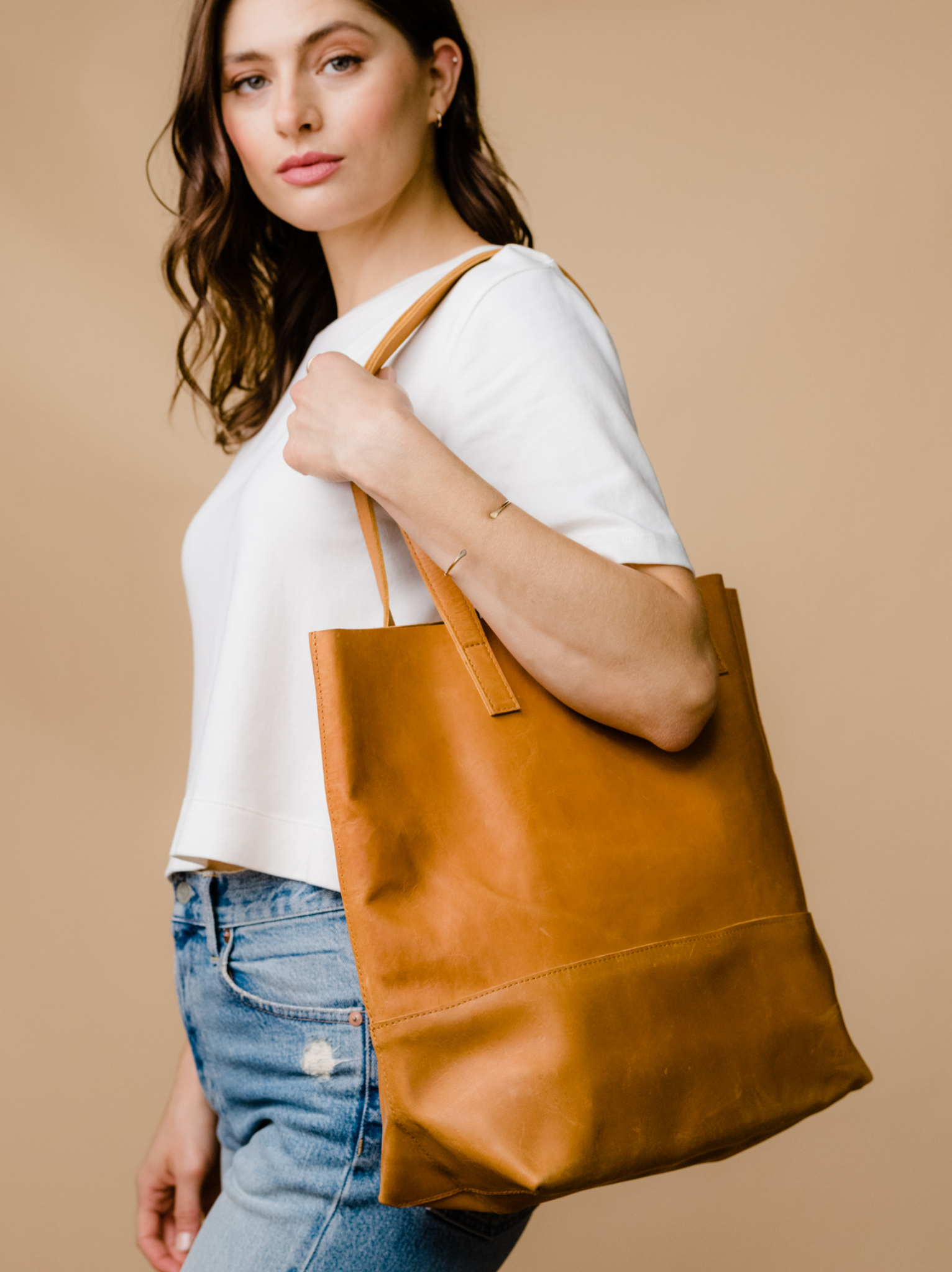 Woman holding an cognac leather tote on her shoulders wearing a white tee with jeans against a neutral background. #Color_Cognac