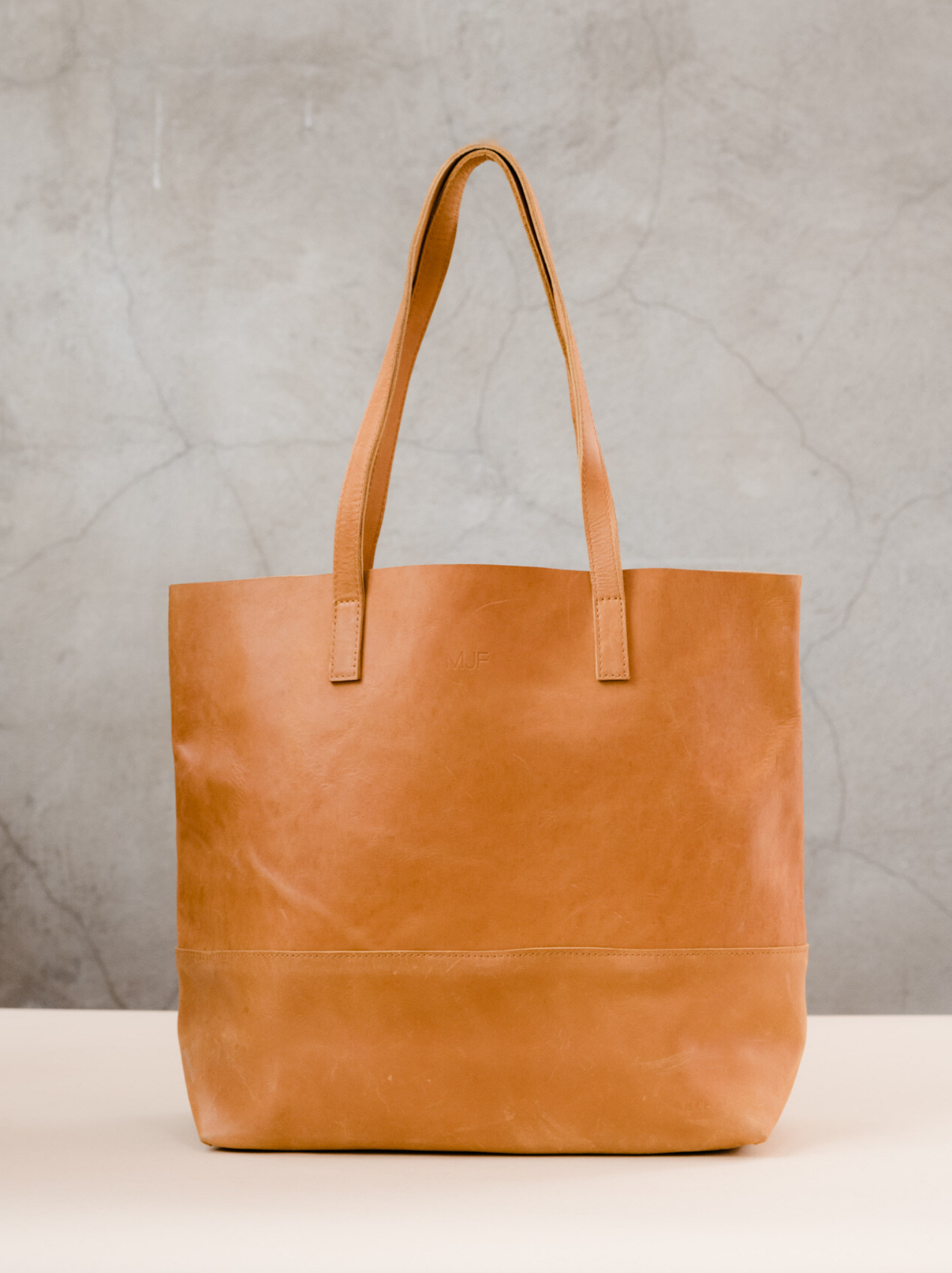 Cognac leather tote bag with two shoulder straps sitting on a tan surface with a neutral background. #Color_Cognac