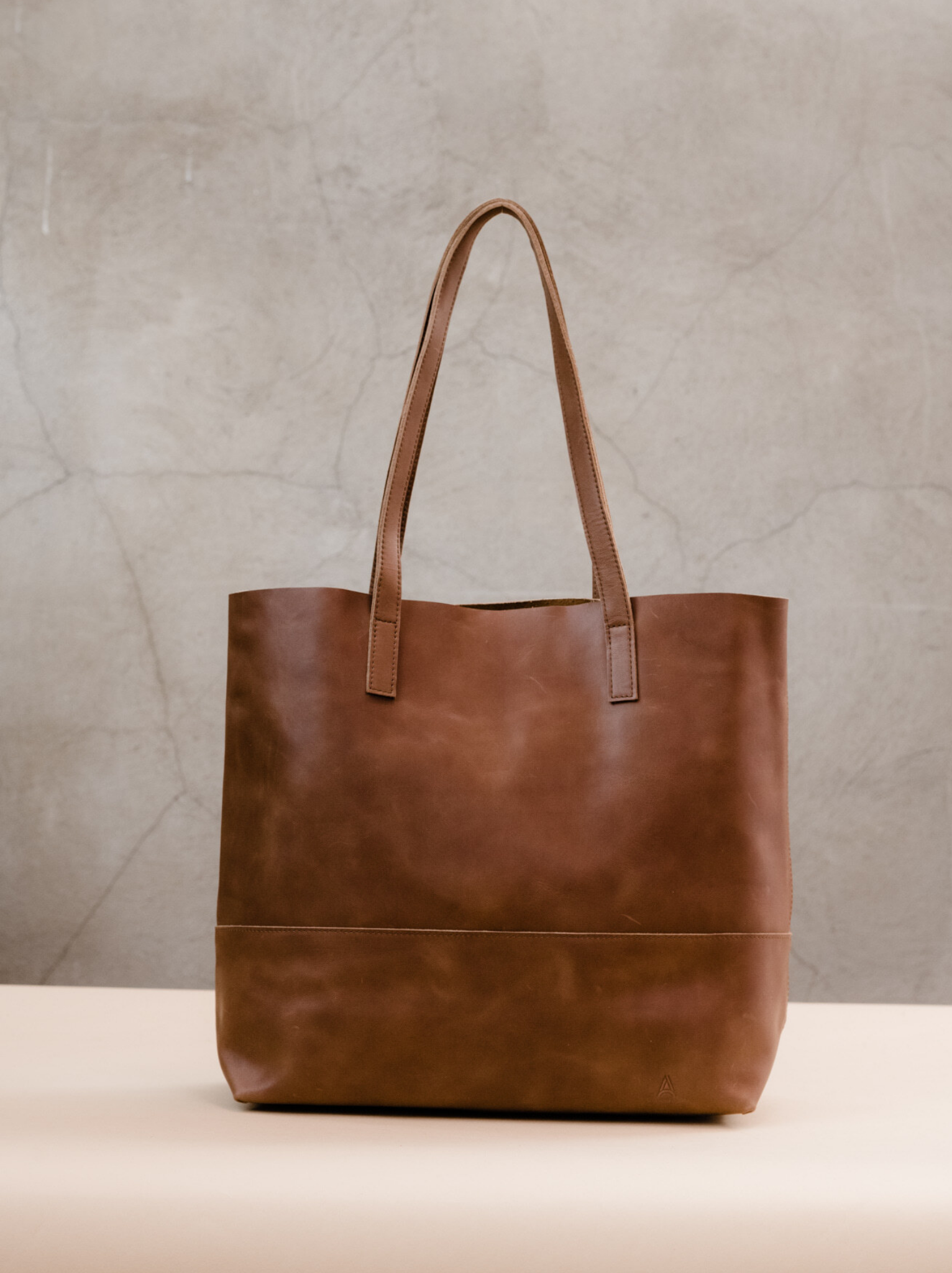 Brown leather tote with two shoulder straps on tan surface against concrete wall. #Color_Whiskey