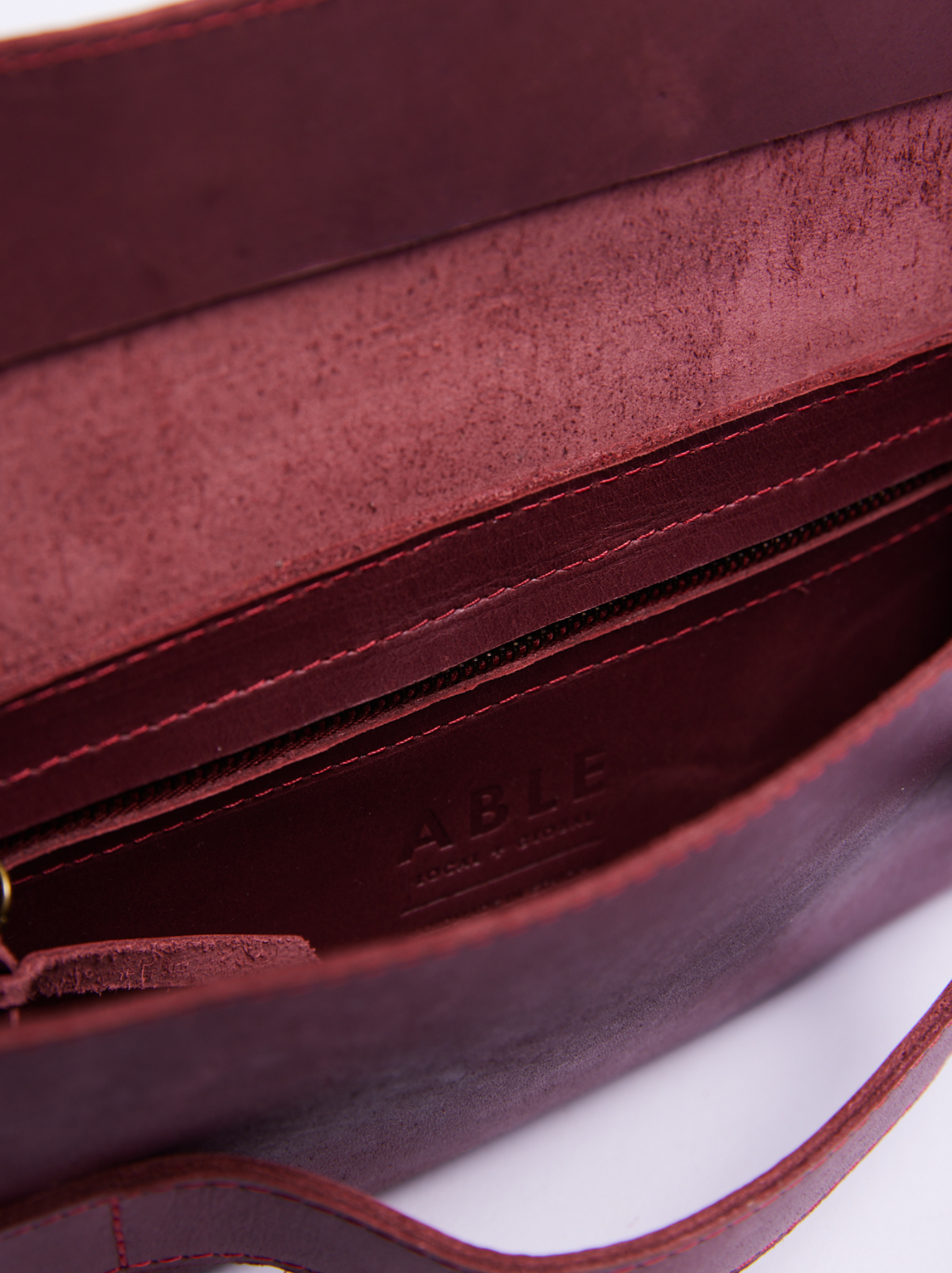 Interior of a burgundy leather clutch bag showing the interior zipper pocket, and the debossed brand name 'ABLE' on the inside. #Color_Wine