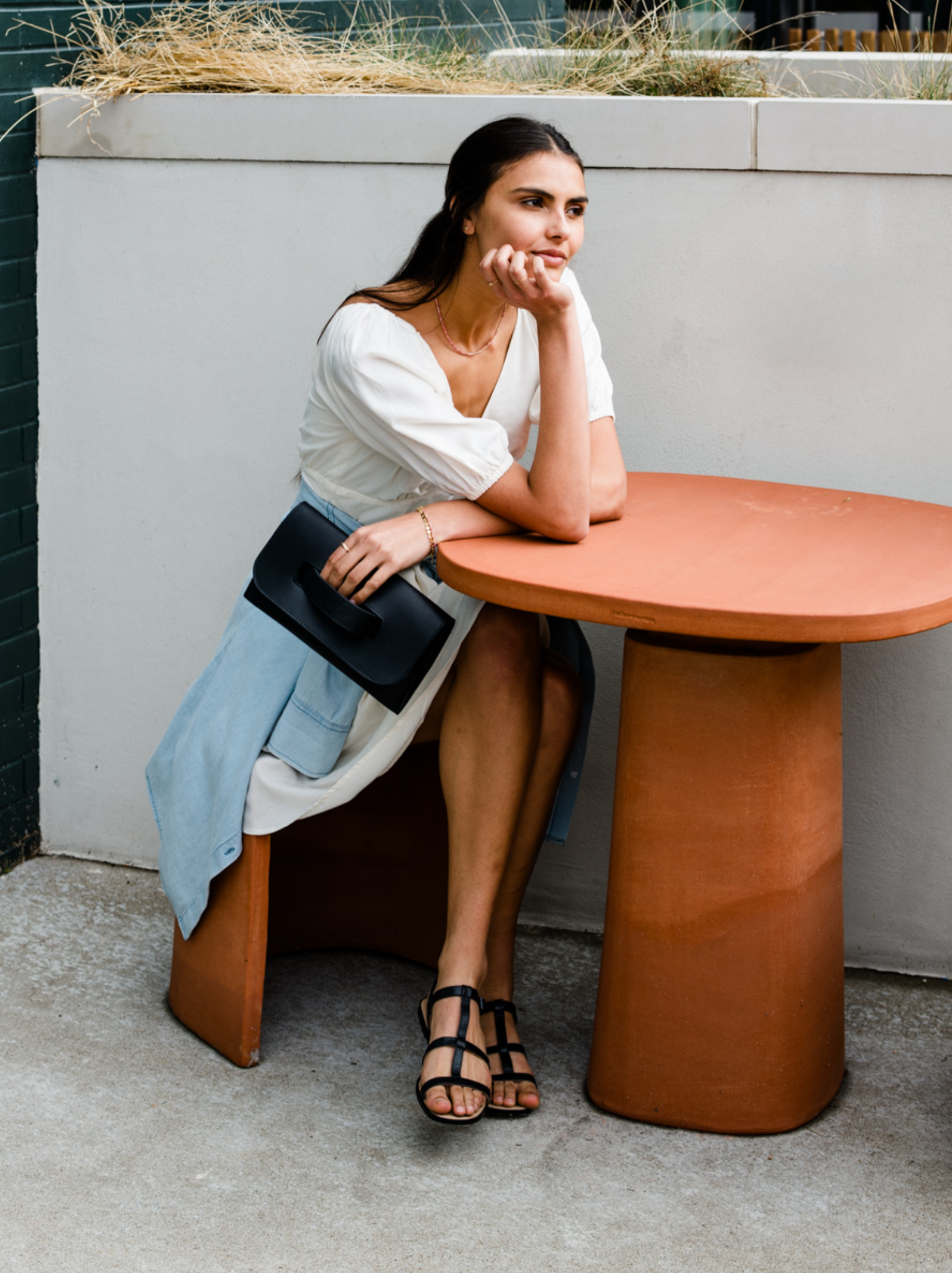 Woman sitting at a round terracotta table outdoors holding a black clutch. #Color_Black