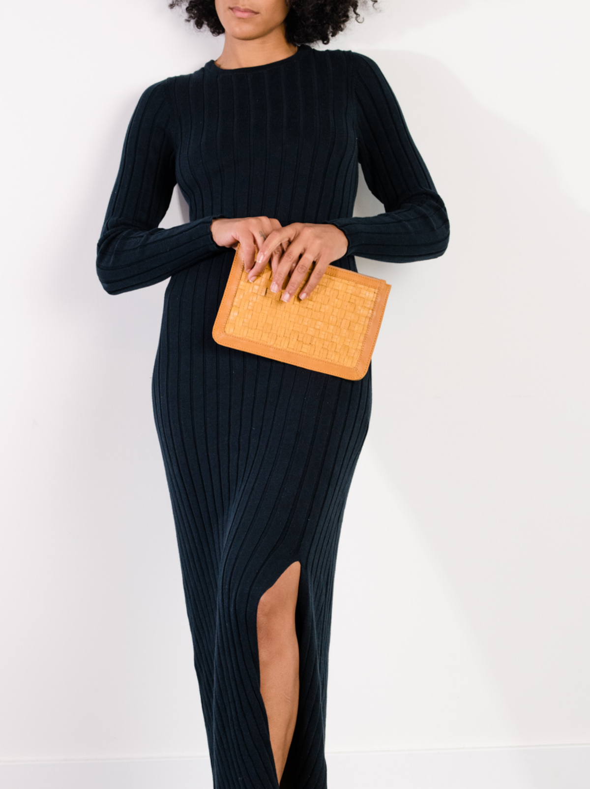 Woman holding a brown basketweave clutch while wearing a black sweater dress against a white wall. #Color_Cognac