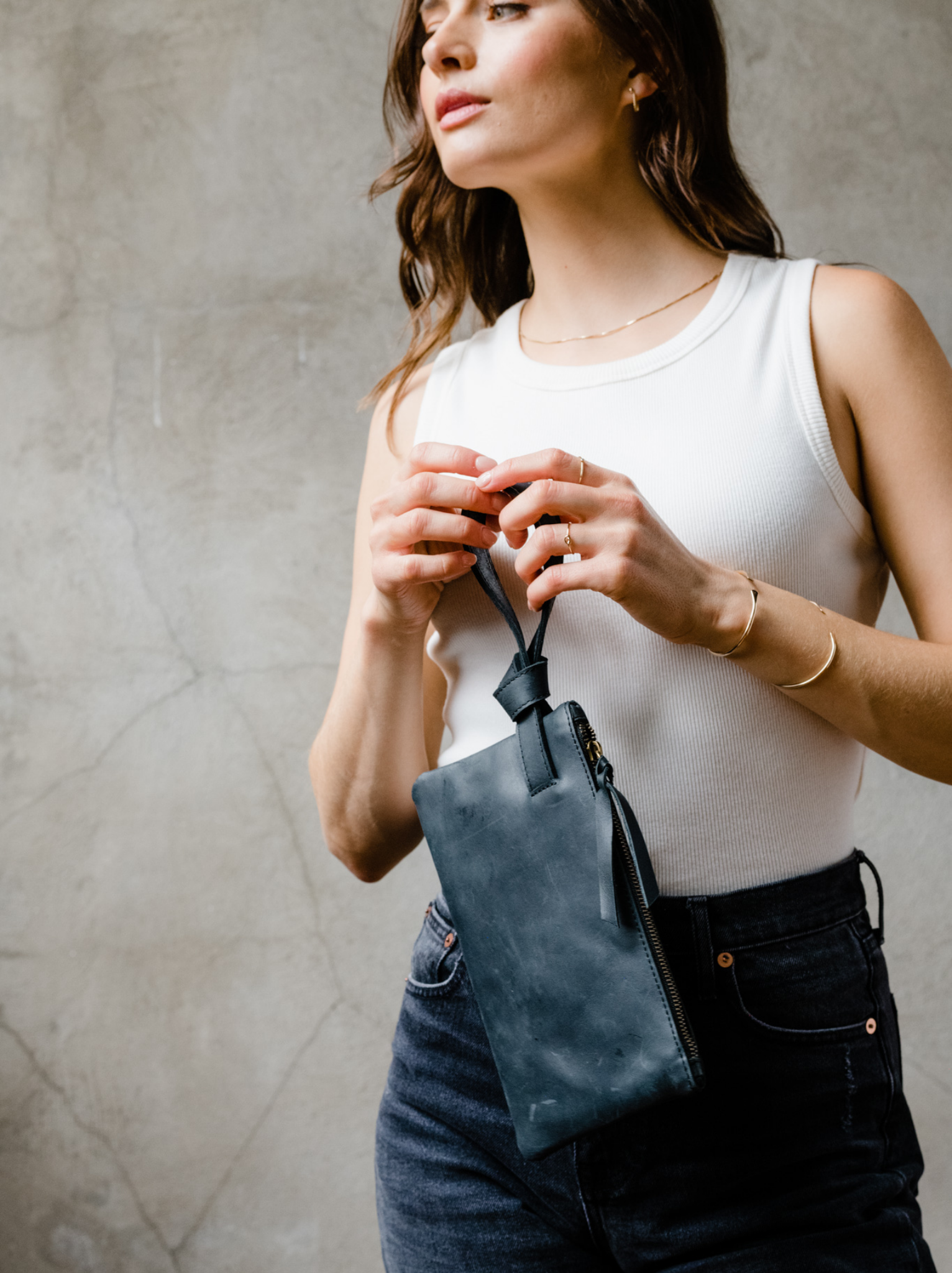 Woman holding a black leather wristlet by the handle in front of her against a neutral background. #Color_Black