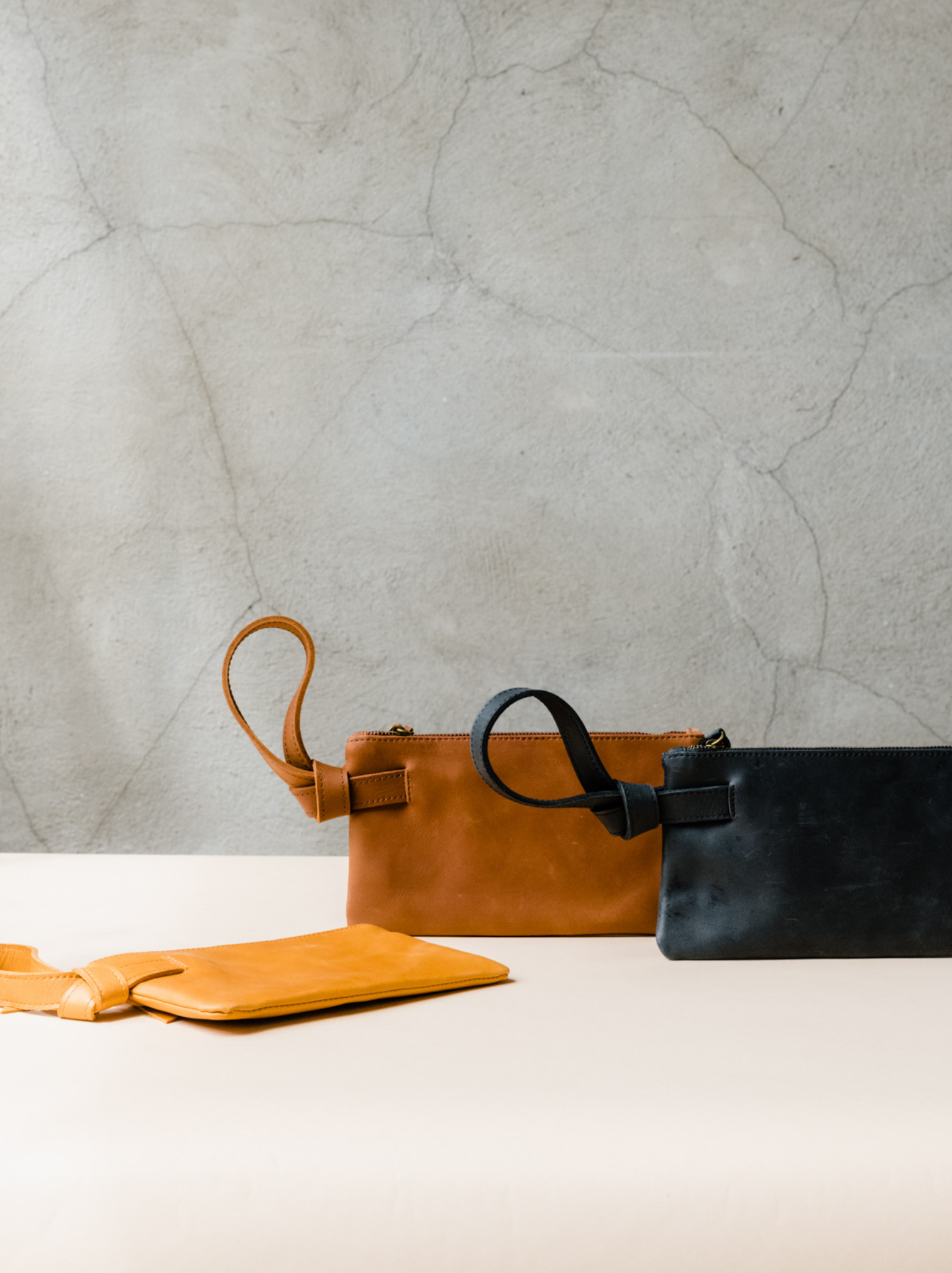 Three leather wristlets, orange, brown, and black, sitting on a tan surface against a neutral background. #Color_Whiskey