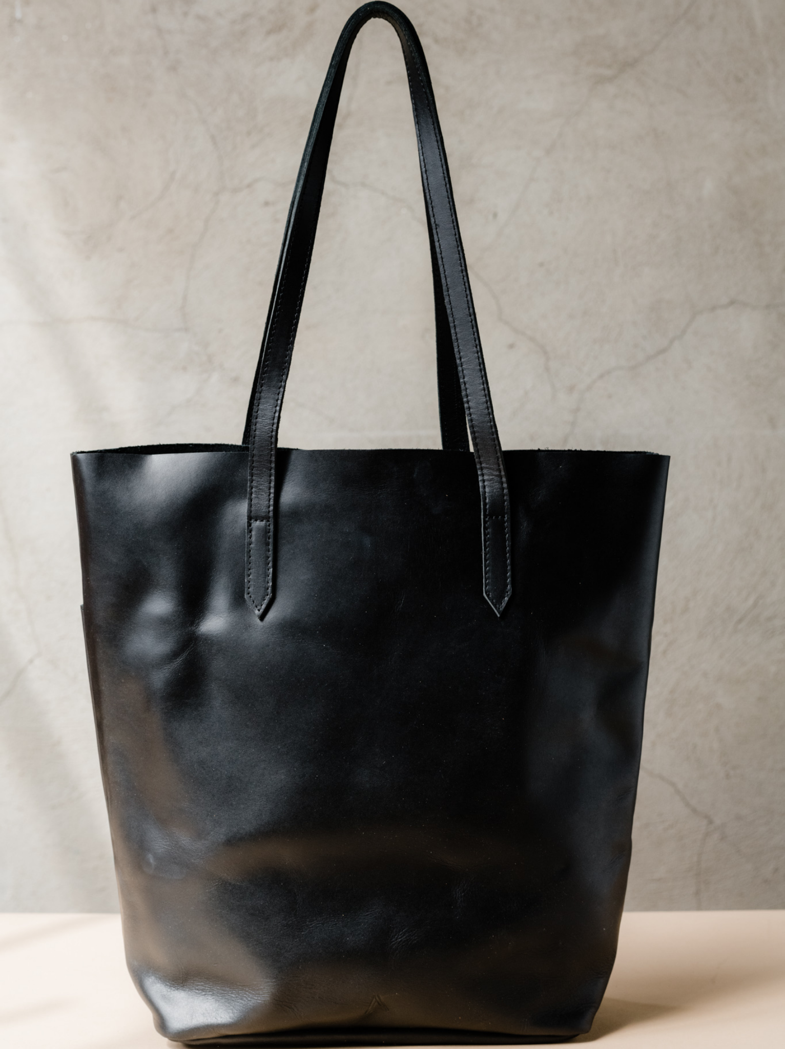 Back of black leather tote with shoulder straps and brand mark at bottom center on tan surface against neutral background. #Color_Black