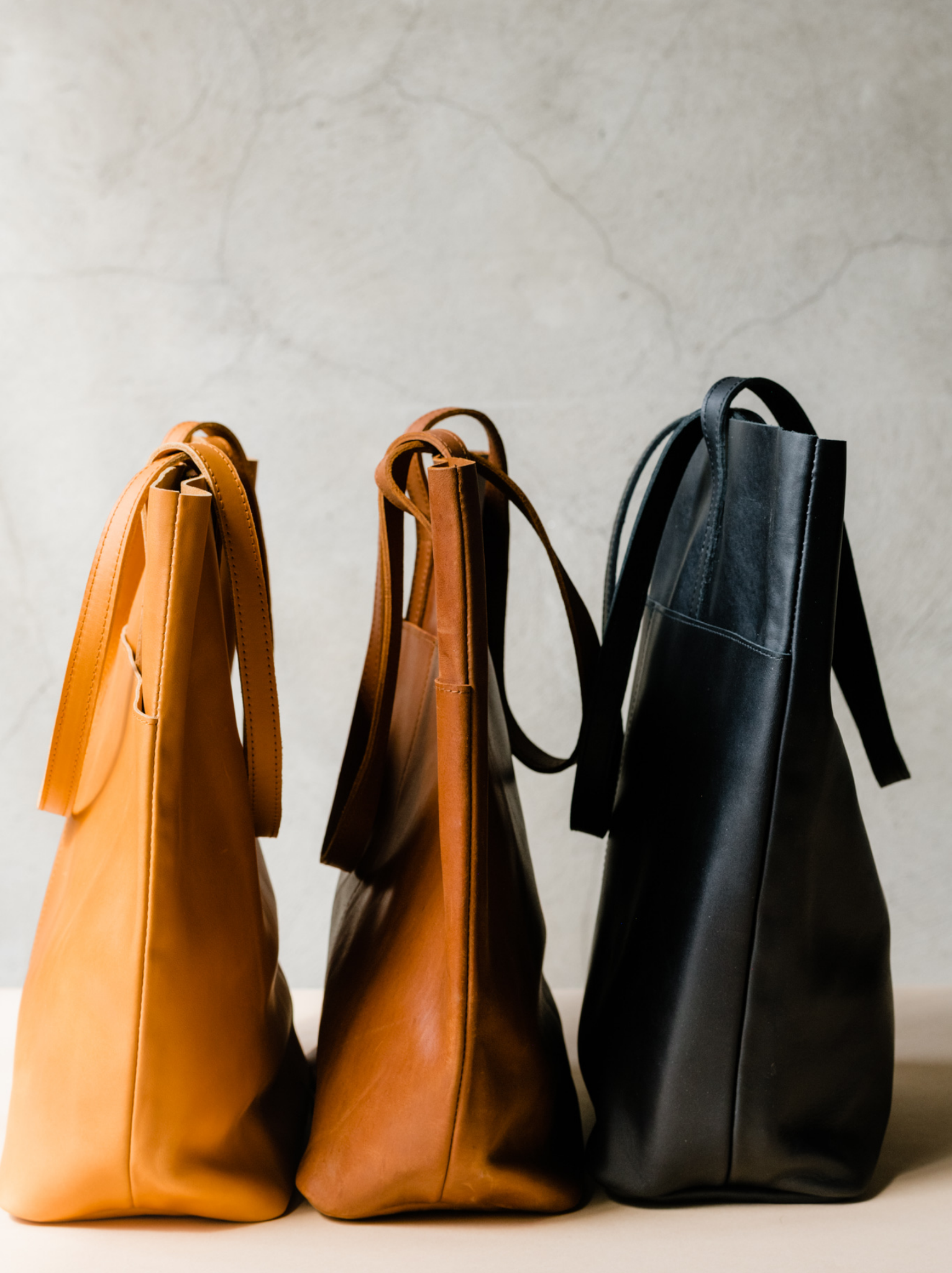 Side view of three leather totes, orange cognac, brown, and black, lined up on a tan surface against a neutral background. #Color_Black