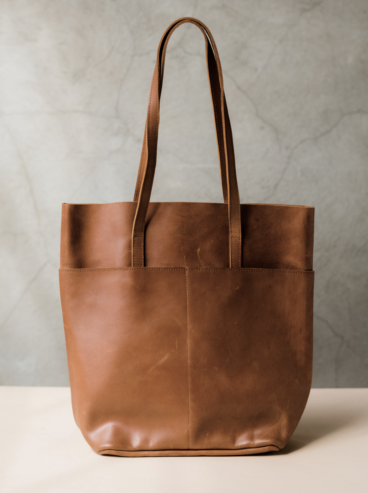 A brown leather tote with two shoulder straps and two slip pockets on a tan surface against a neutral background. #Color_Whiskey
