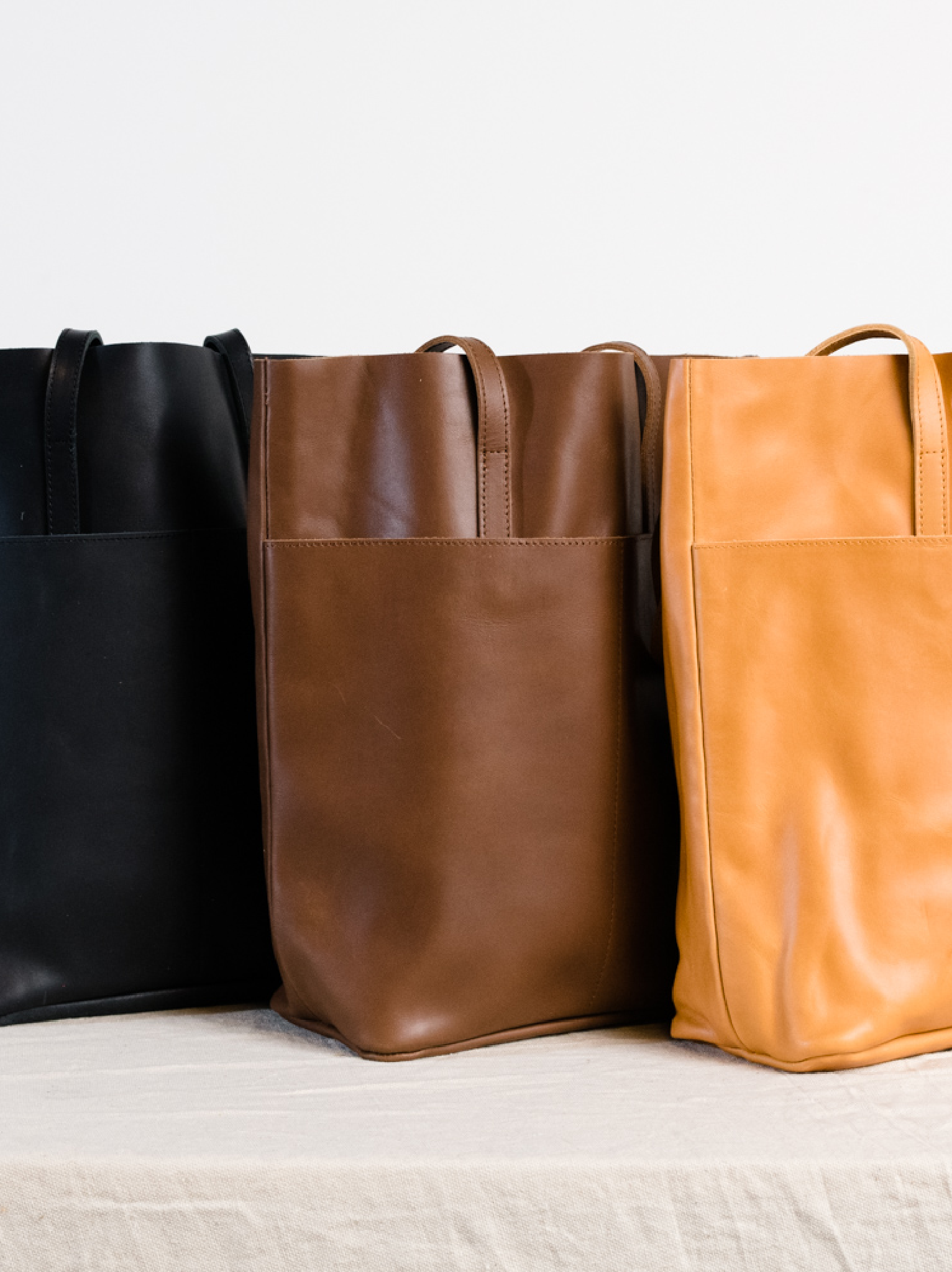 Three leather totes, black, brown, and orange cognac, lined up on a tan surface against a white background. #Color_Whiskey