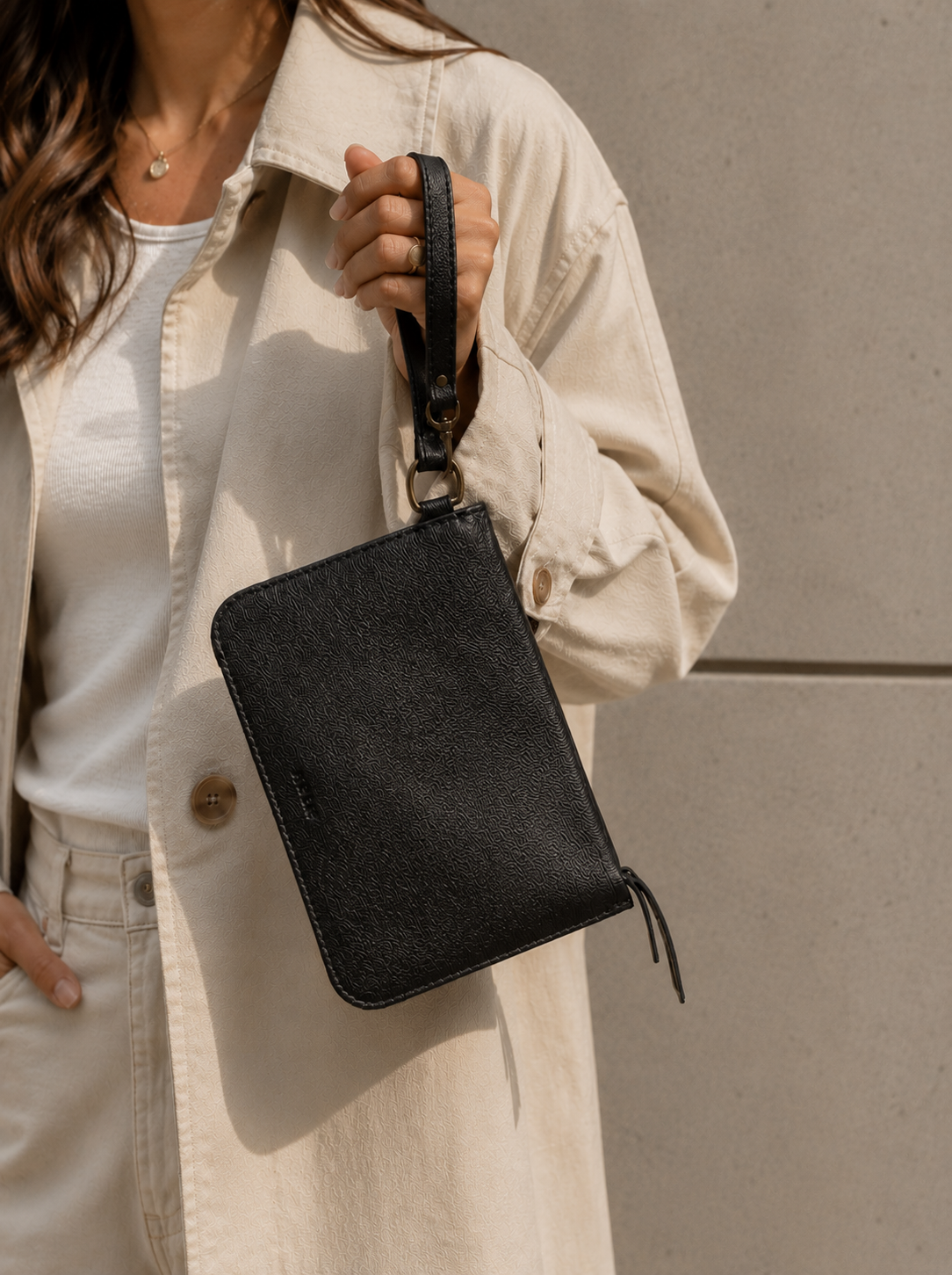 Woman holding a pebbled textured black leather pouch by its leather wristlet strap against a neutral background. #Color_Pebbled Black