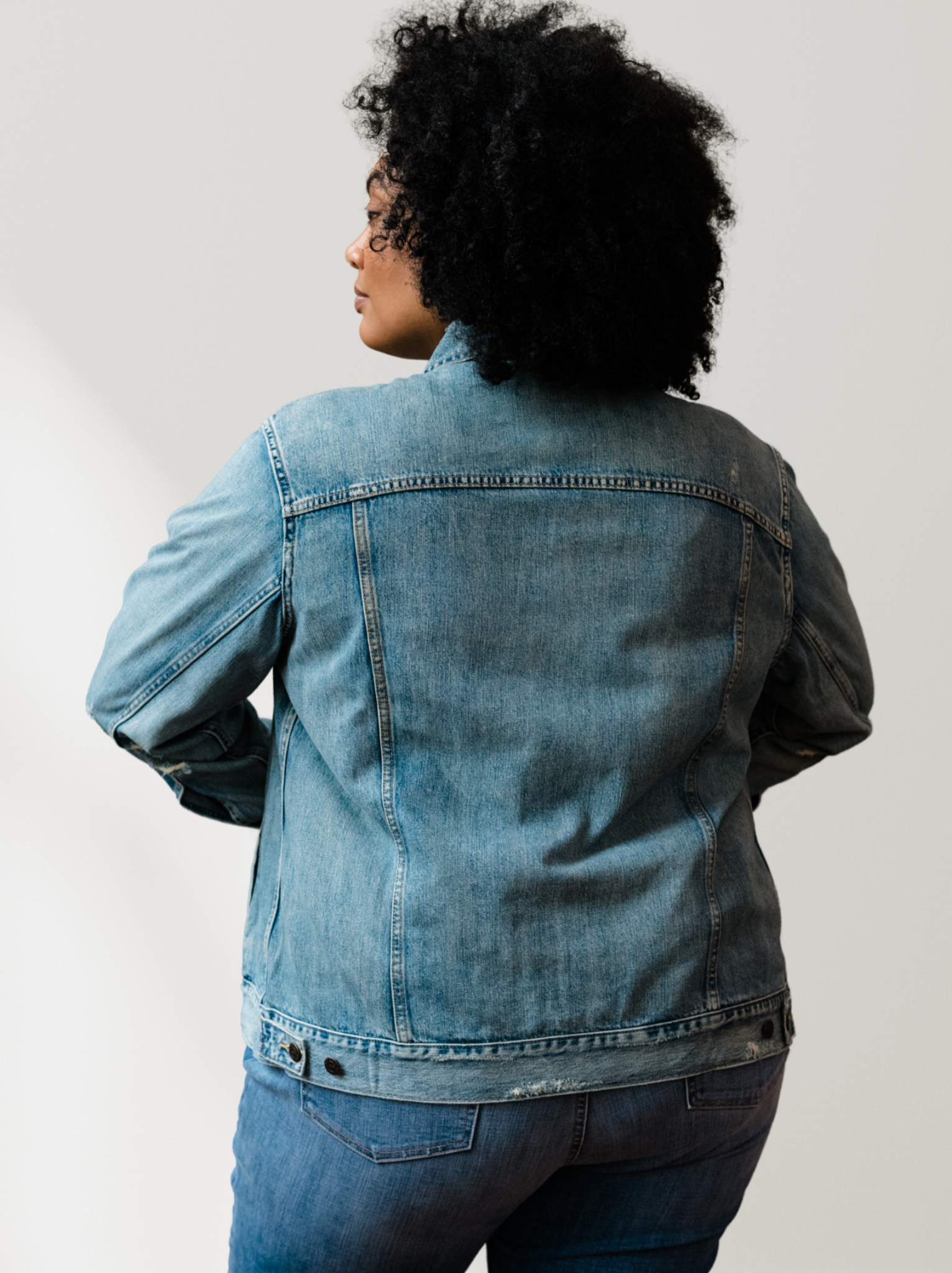 Back of woman wearing a light blue denim jacket with light distressing and jeans against a neutral background. #Color_Original Wash