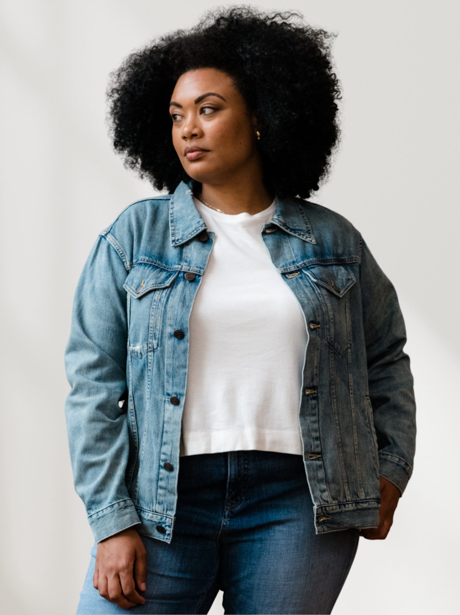 A woman wearing a light blue denim jacket with light distressing, front pockets and button front over a white shirt with denim jeans against a white background. #Color_Original Wash