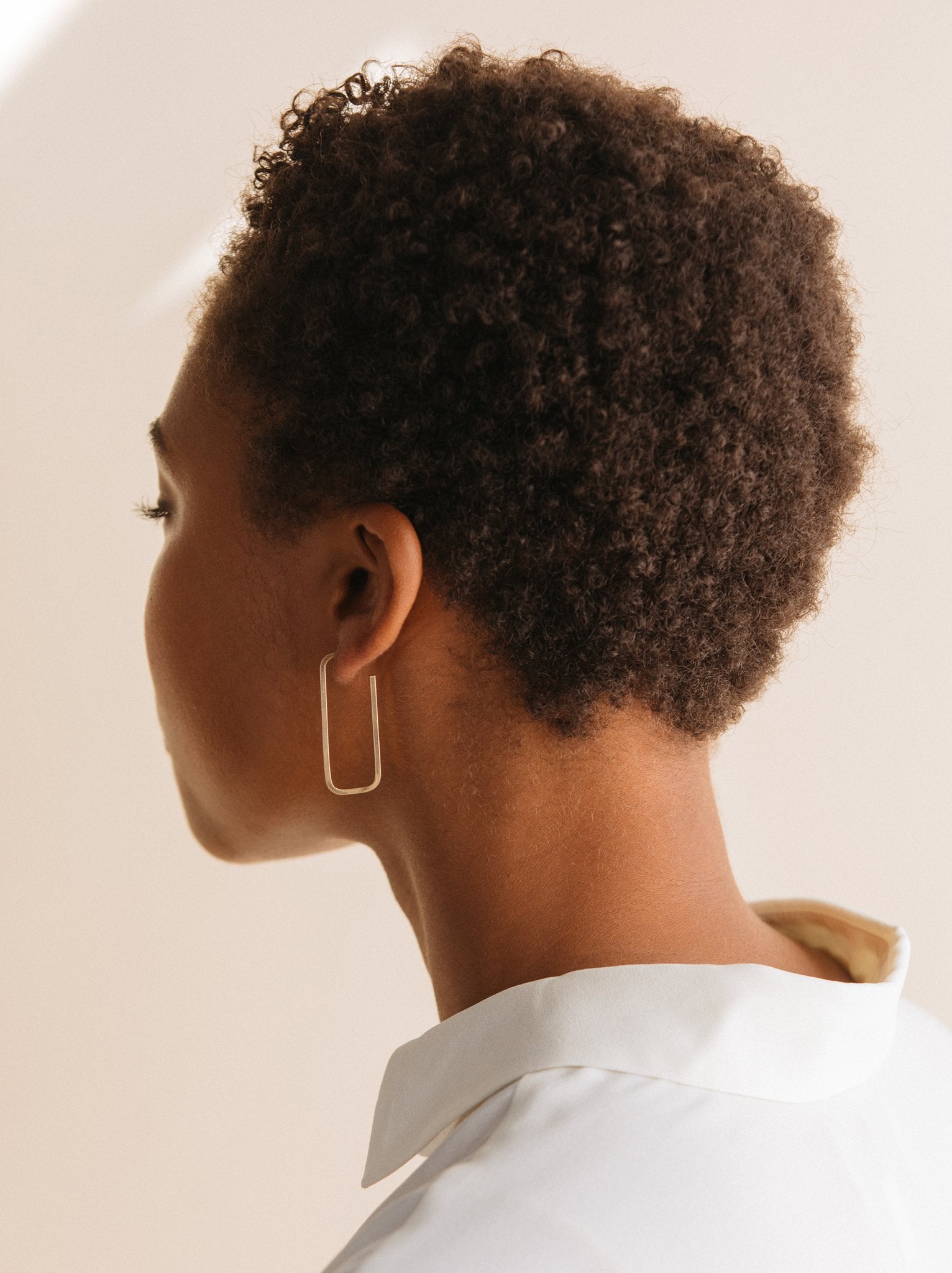 A woman wearing gold rectangle style hoop earrings against a neutral background.