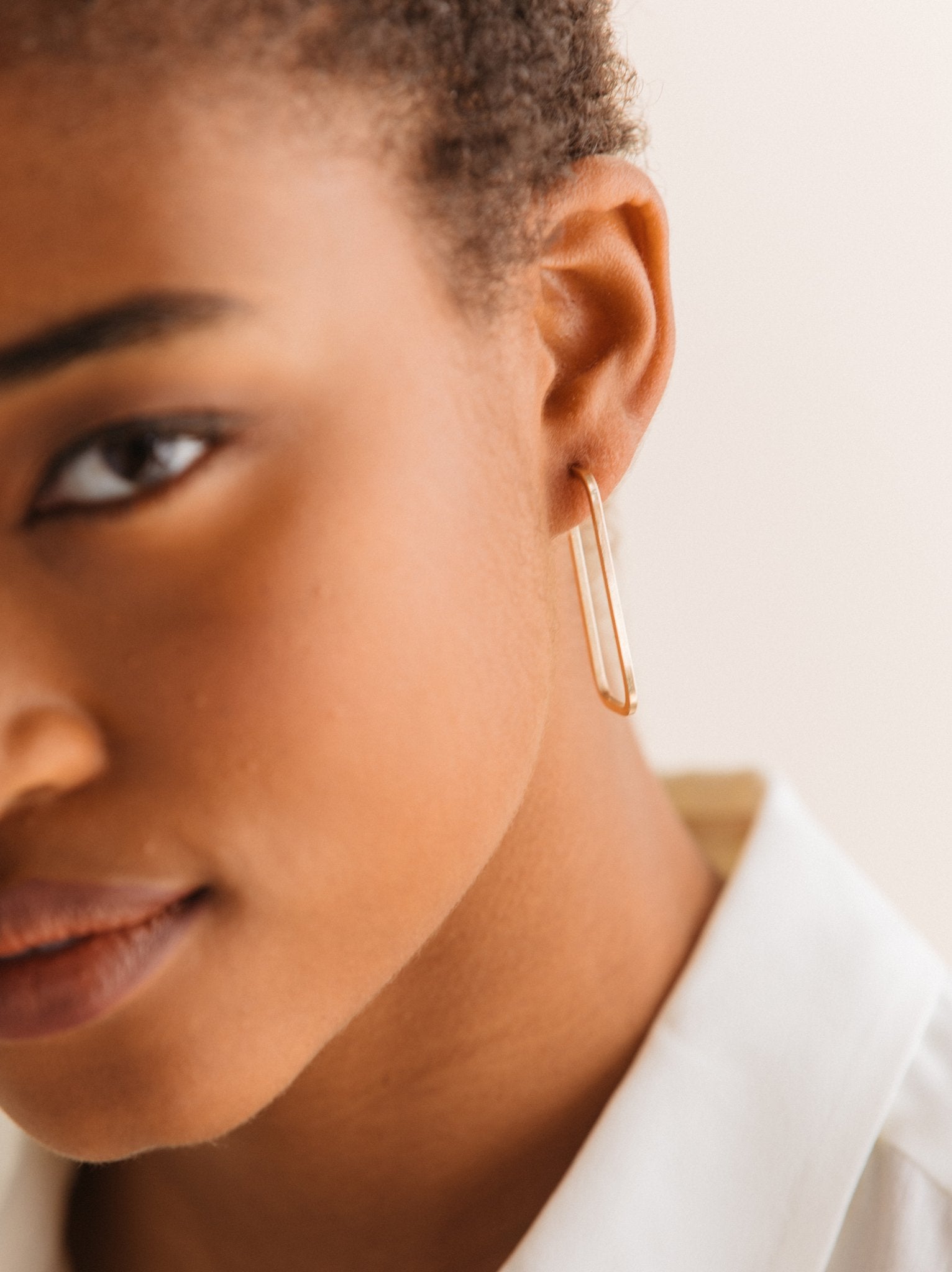 A woman wearing gold rectangle style hoop earrings showing the thickness against a neutral background.