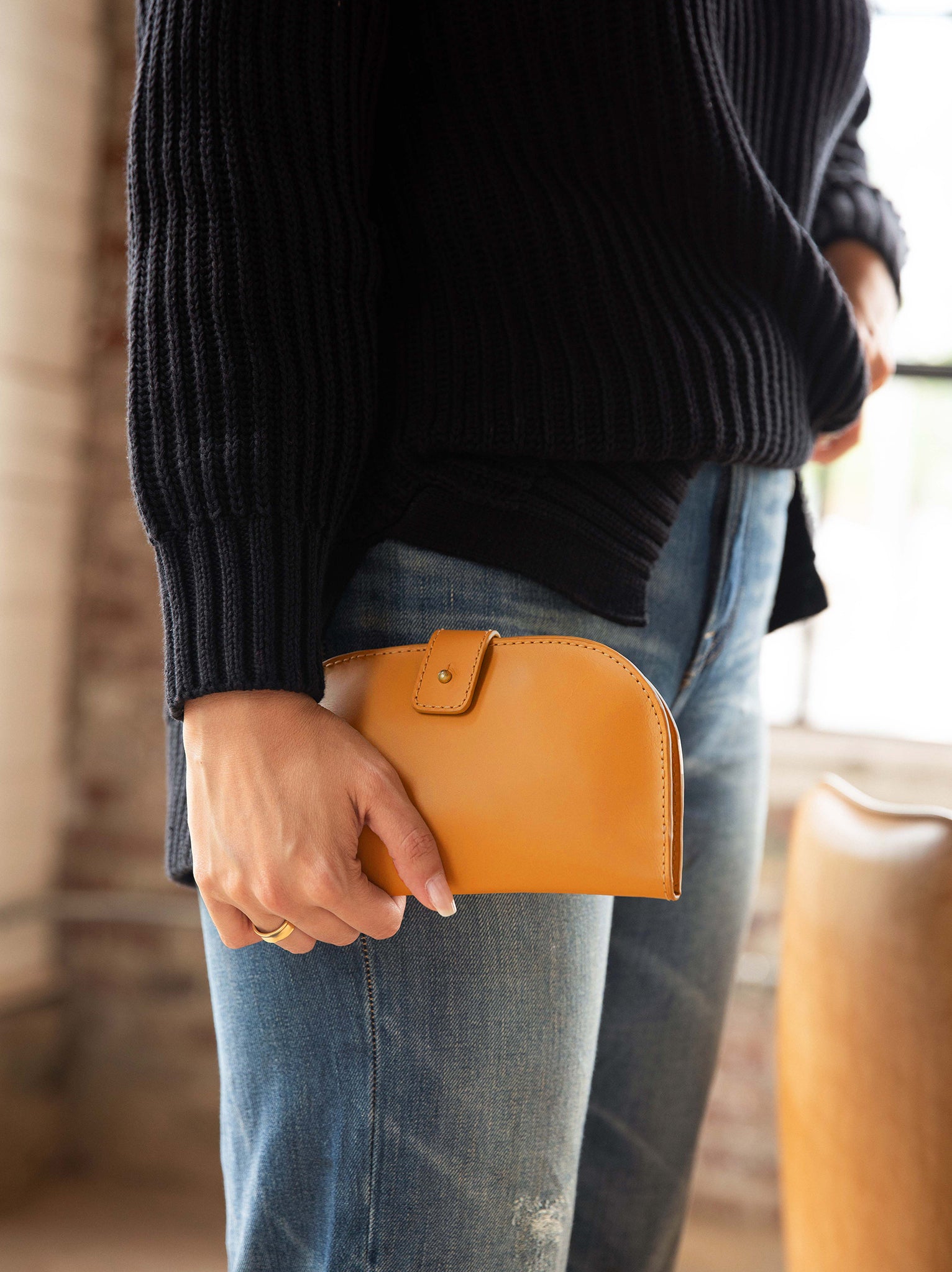 Woman holding Cognac bifold wallet in her hand wearing jeans and a sweater in a room with a brick wall. #Color_Cognac