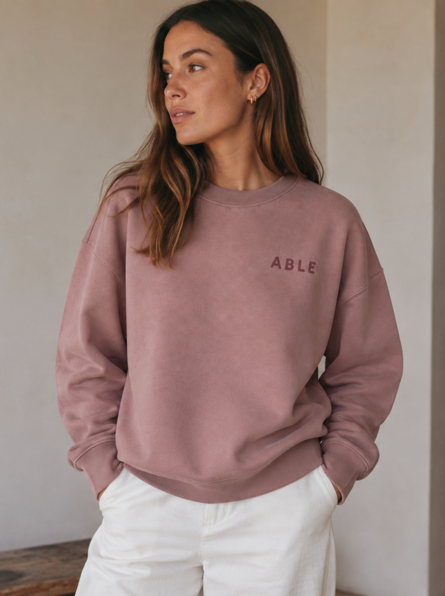 A woman wearing a dusty pink crewneck sweatshirt with the word 'ABLE' printed in dark pink on the left chest, paired with white pants, standing against a neutral beige wall. #Color_Hazel Pink