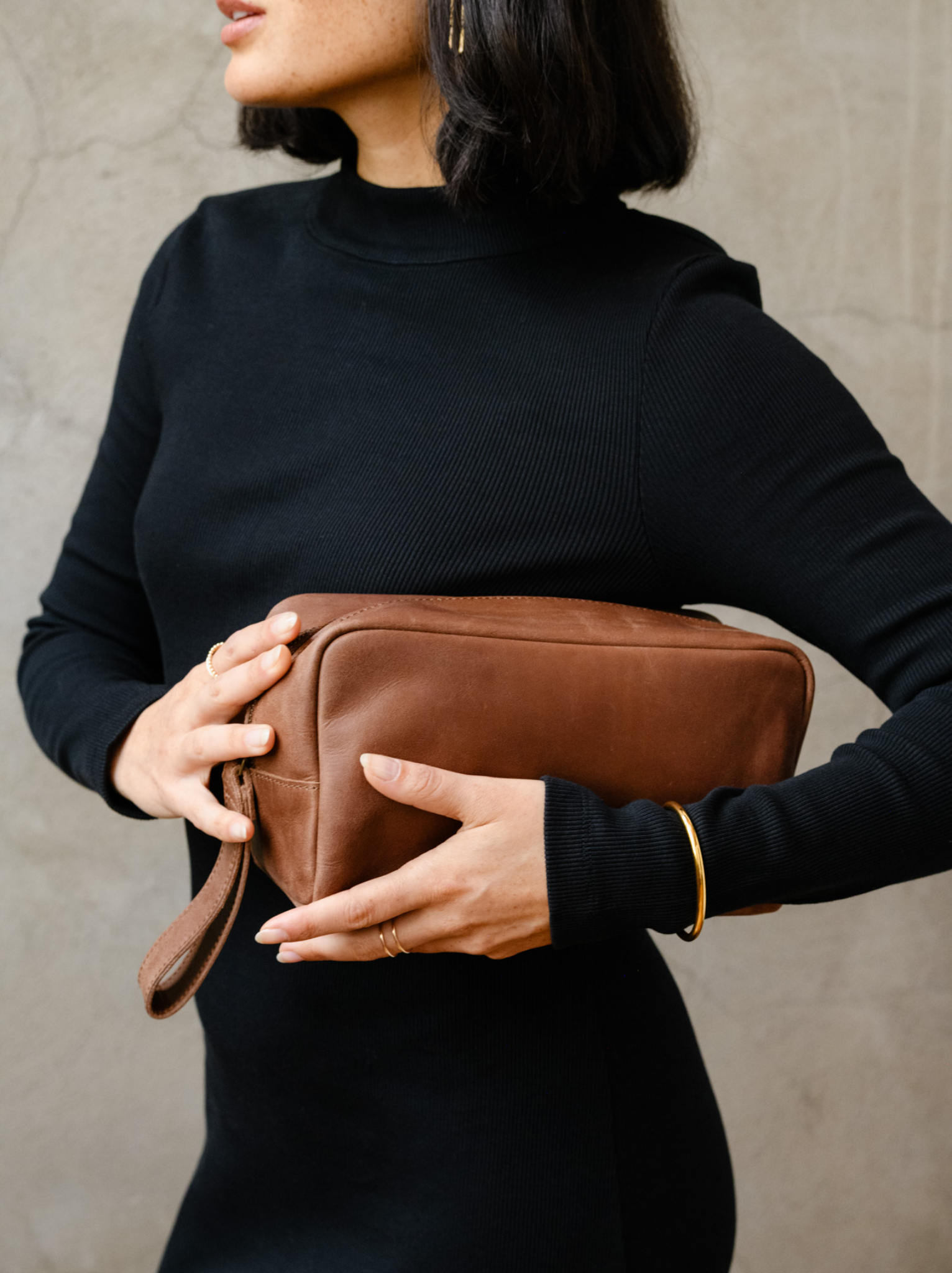 A woman holding a brown leather travel case with a leather hang strap. #Color_Whiskey