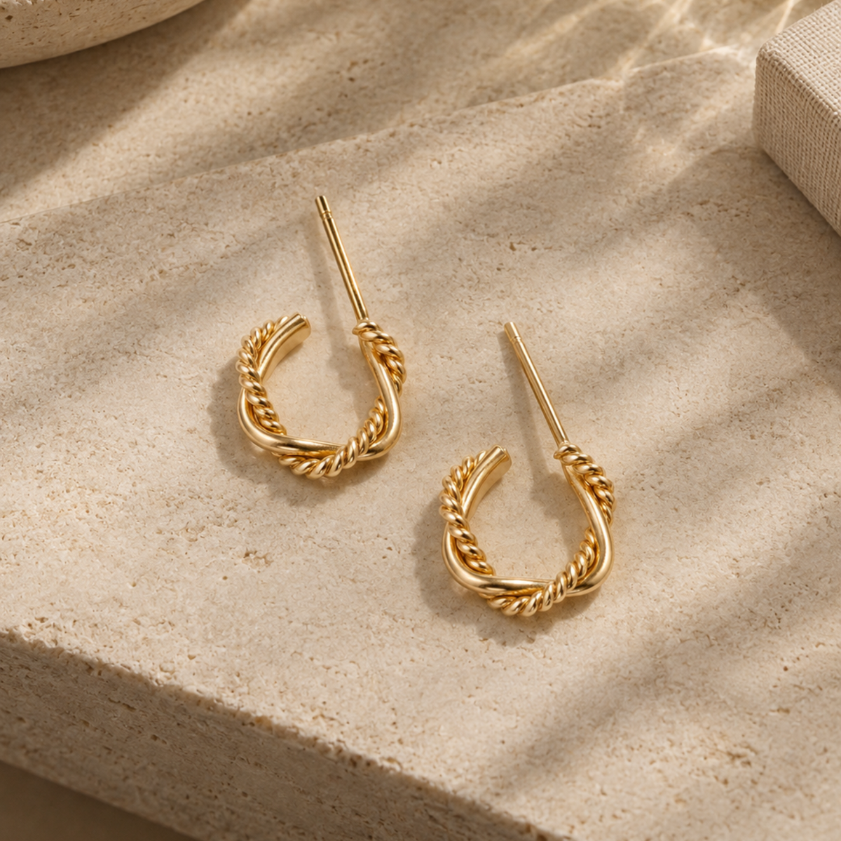 A pair of small gold twisted design hoop earrings on a stone slab.