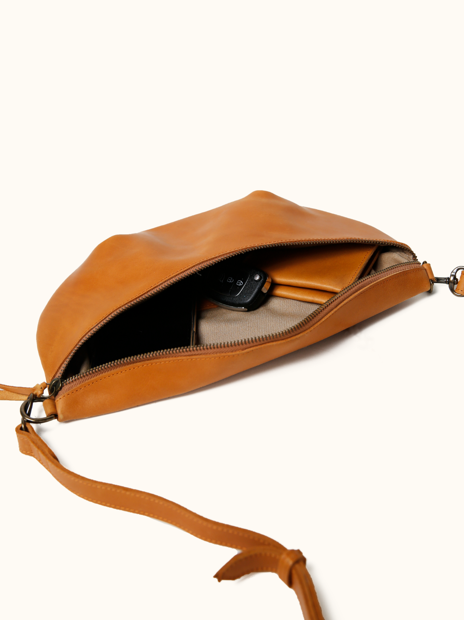 Cognac leather belt bag open showing lined interior with keys, wallet, and phone inside on white background. #Color_Cognac