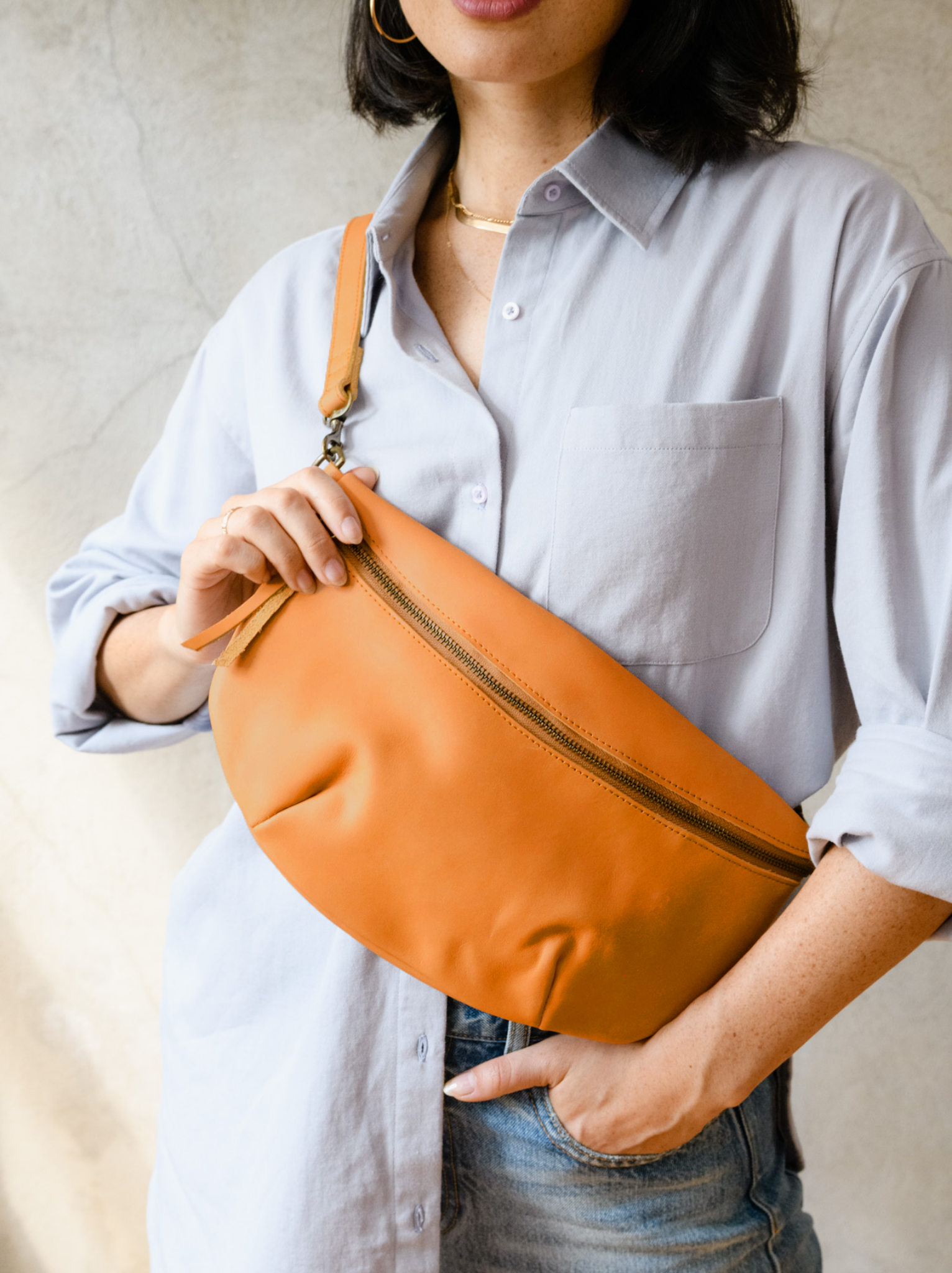 Woman wearing a cognac zip up leather belt bag across her chest with blue button down shirt and jeans against concrete wall. #Color_Cognac