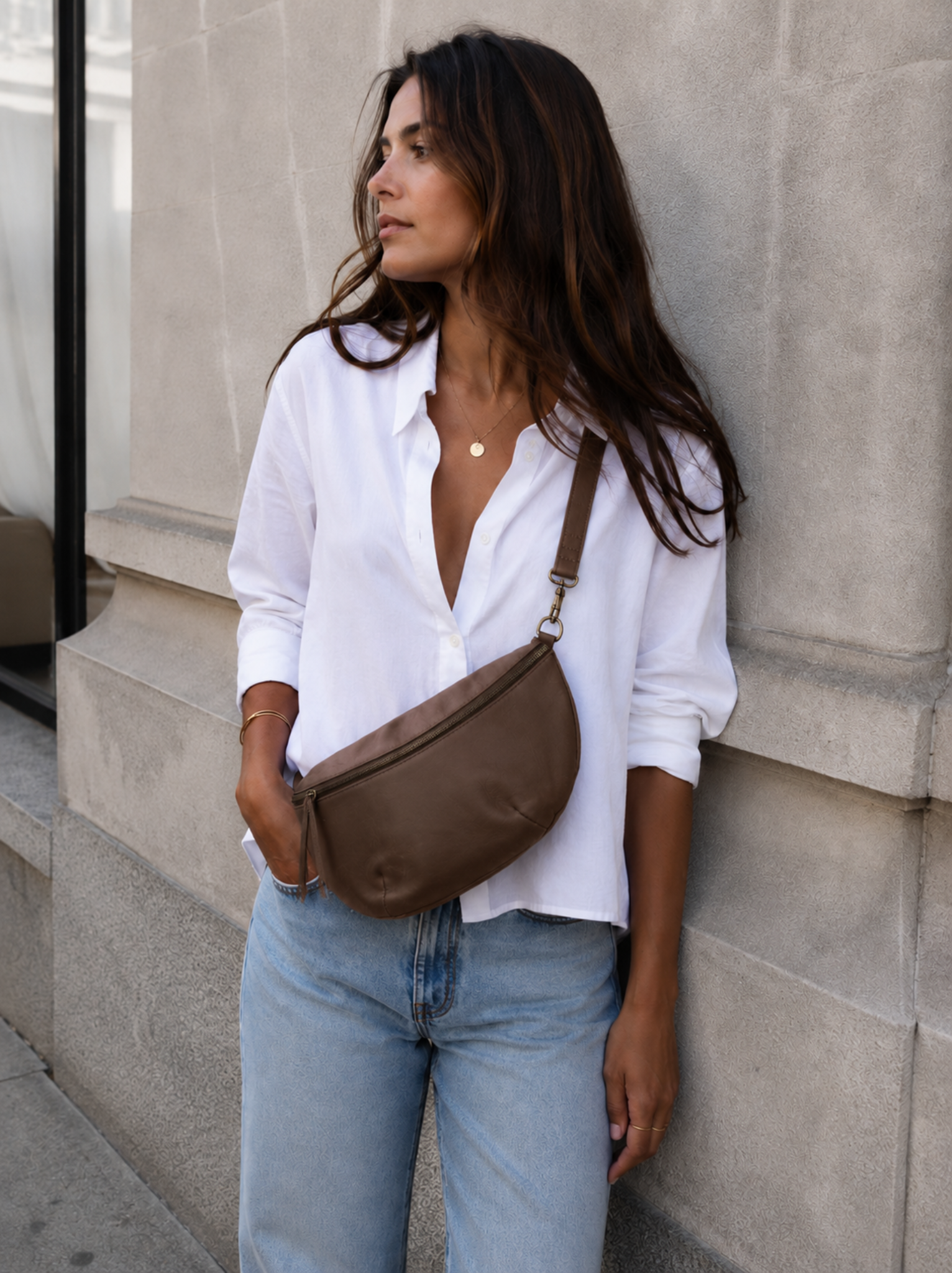 A woman wearing a white button-down shirt and light blue jeans carries a brown leather crescent-shaped crossbody bag against a light gray stone wall. #Color_Whiskey