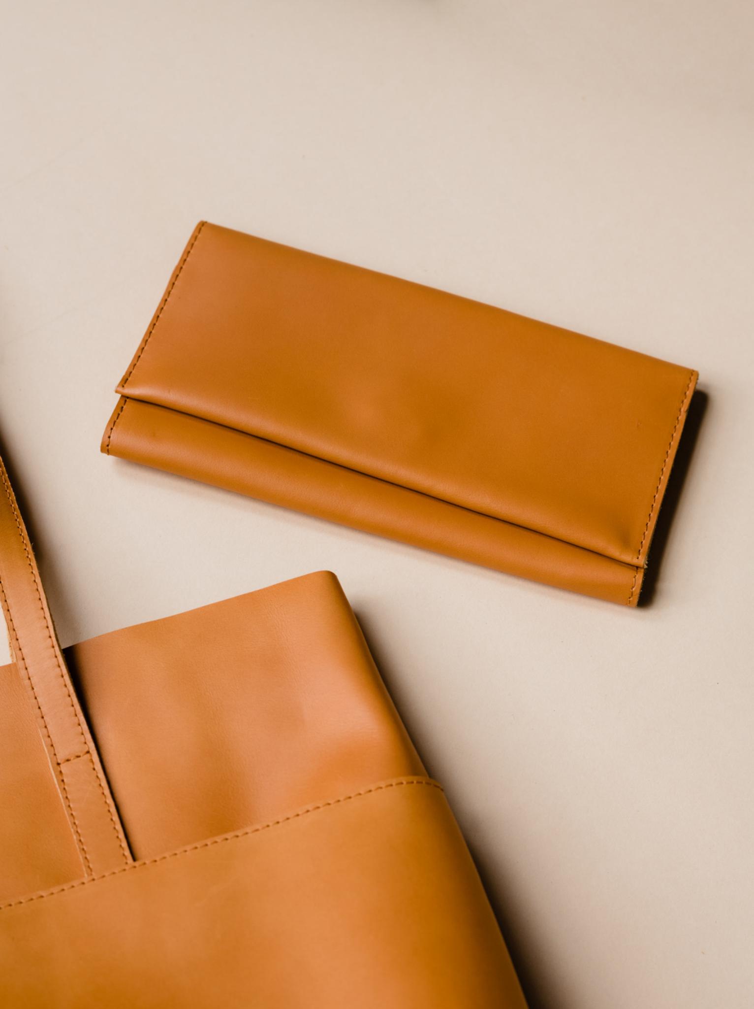 Orange cognac leather wallet lying next to leather tote on neutral background. #Color_Cognac