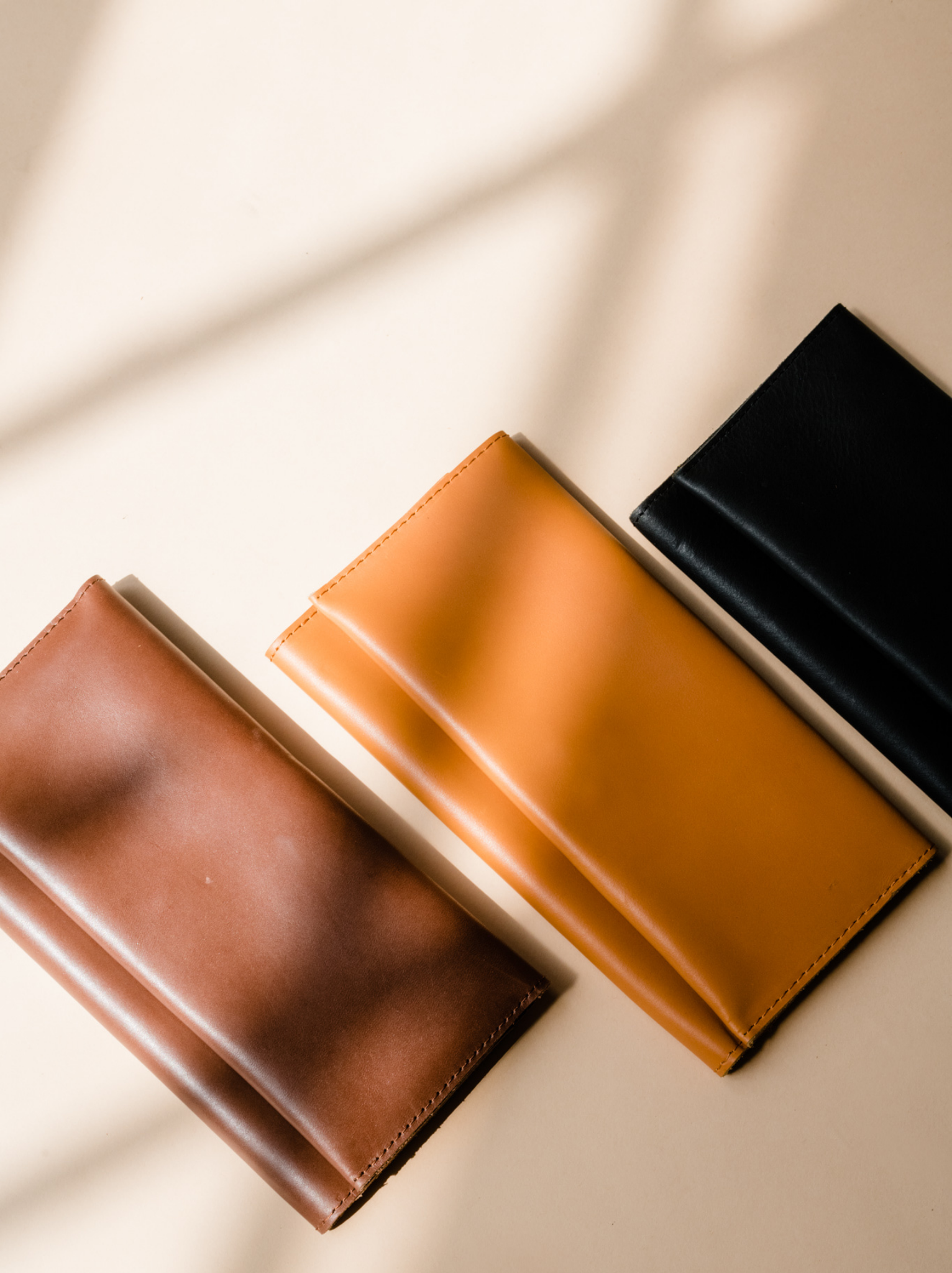Three leather wallets, brown, orange cognac, and black, lying next to each other on neutral background. #Color_Black